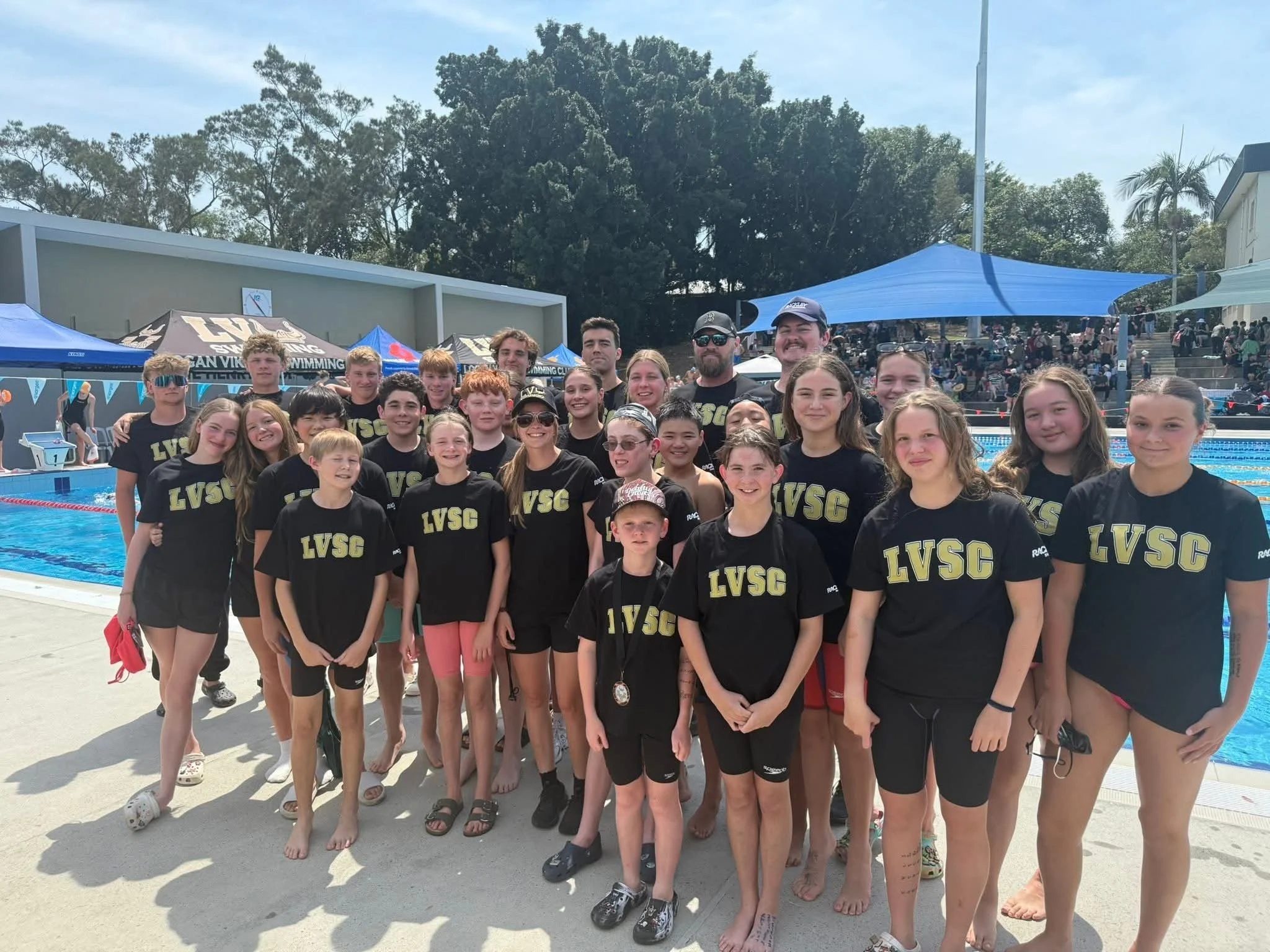 Swimmers and coaches posing for a group photo at an outdoor swimming pool during a swim meet.