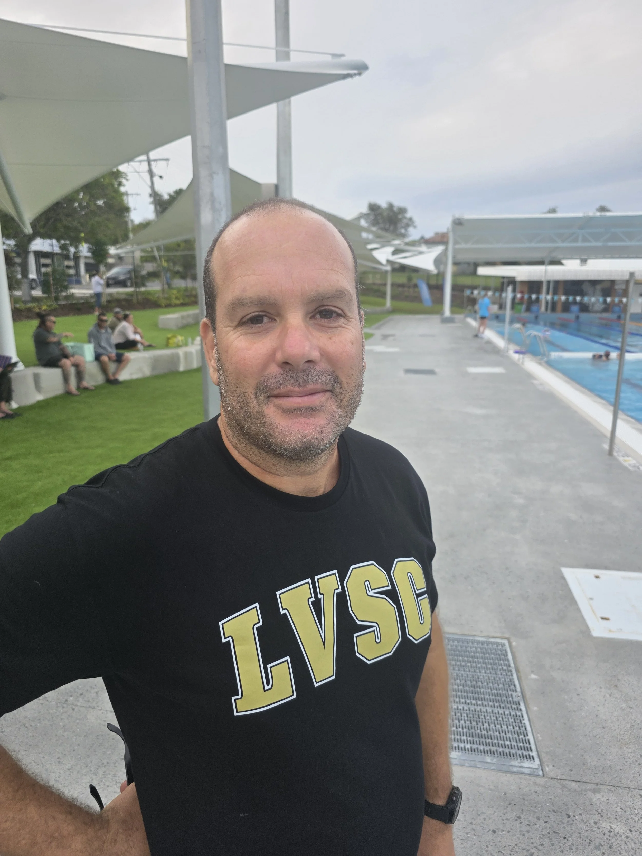 A man wearing a black T-shirt with gold and white text standing outside near a swimming pool with shade sails and other people in the background.