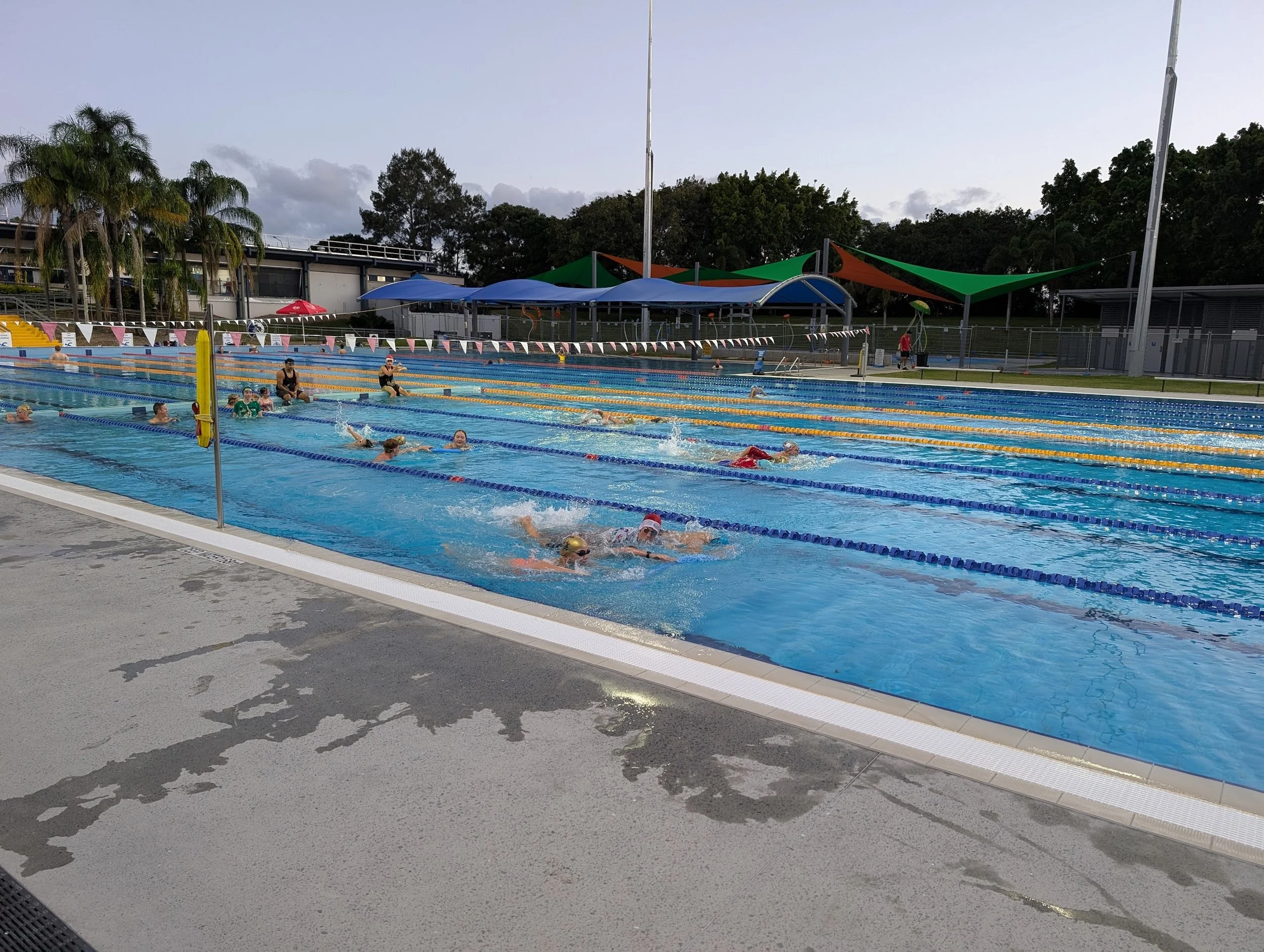 Outdoor swimming pool with multiple lanes, children swimming, parents supervising, shaded structures, and trees in the background.