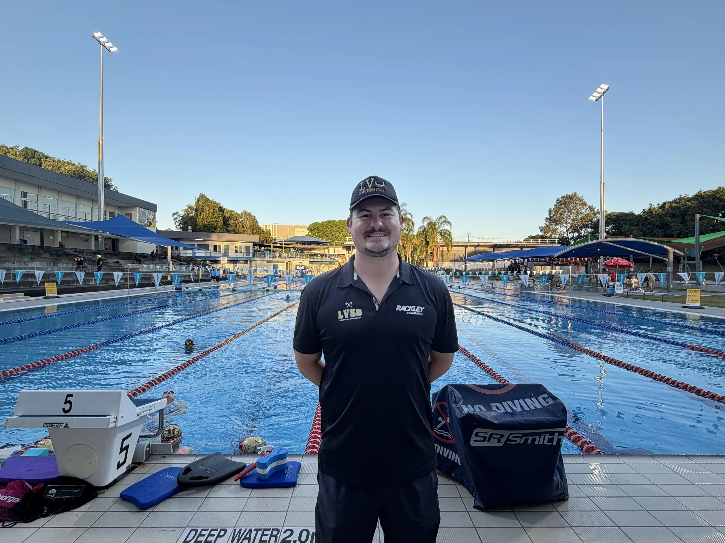 A man standing at the side of an outdoor swimming pool, smiling at the camera, with swimmers in lanes in the background. It is late afternoon with a clear sky. The man is wearing a dark polo shirt and a dark cap, and there are swimming gear and equipment on the tiled pool deck.