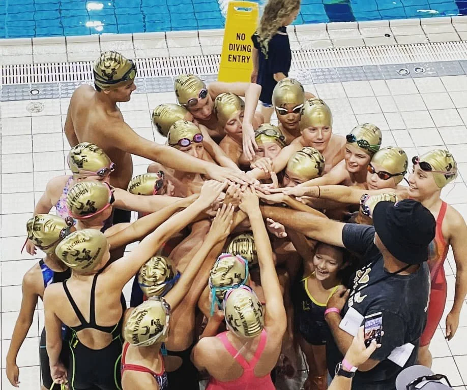 Swimmers in a circle at the poolside, wearing gold swim caps and goggles, with their hands layered together in a team cheer, and a coach or adult recording the moment.