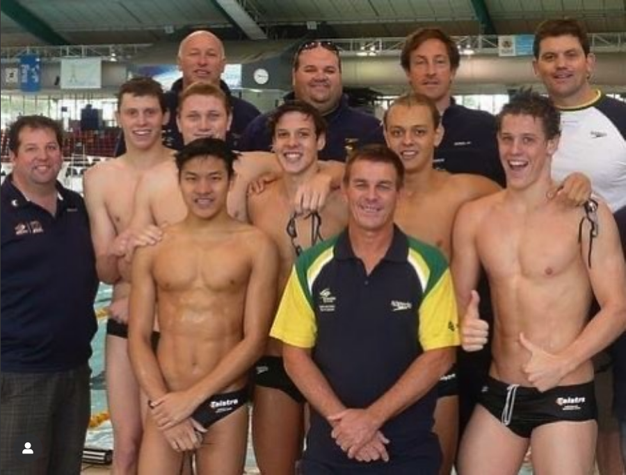 Group of male water polo players and coaches posing together at an indoor swimming pool. Some players are shirtless, wearing swimsuits, and holding water polo caps. Everyone is smiling and showing positive energy.