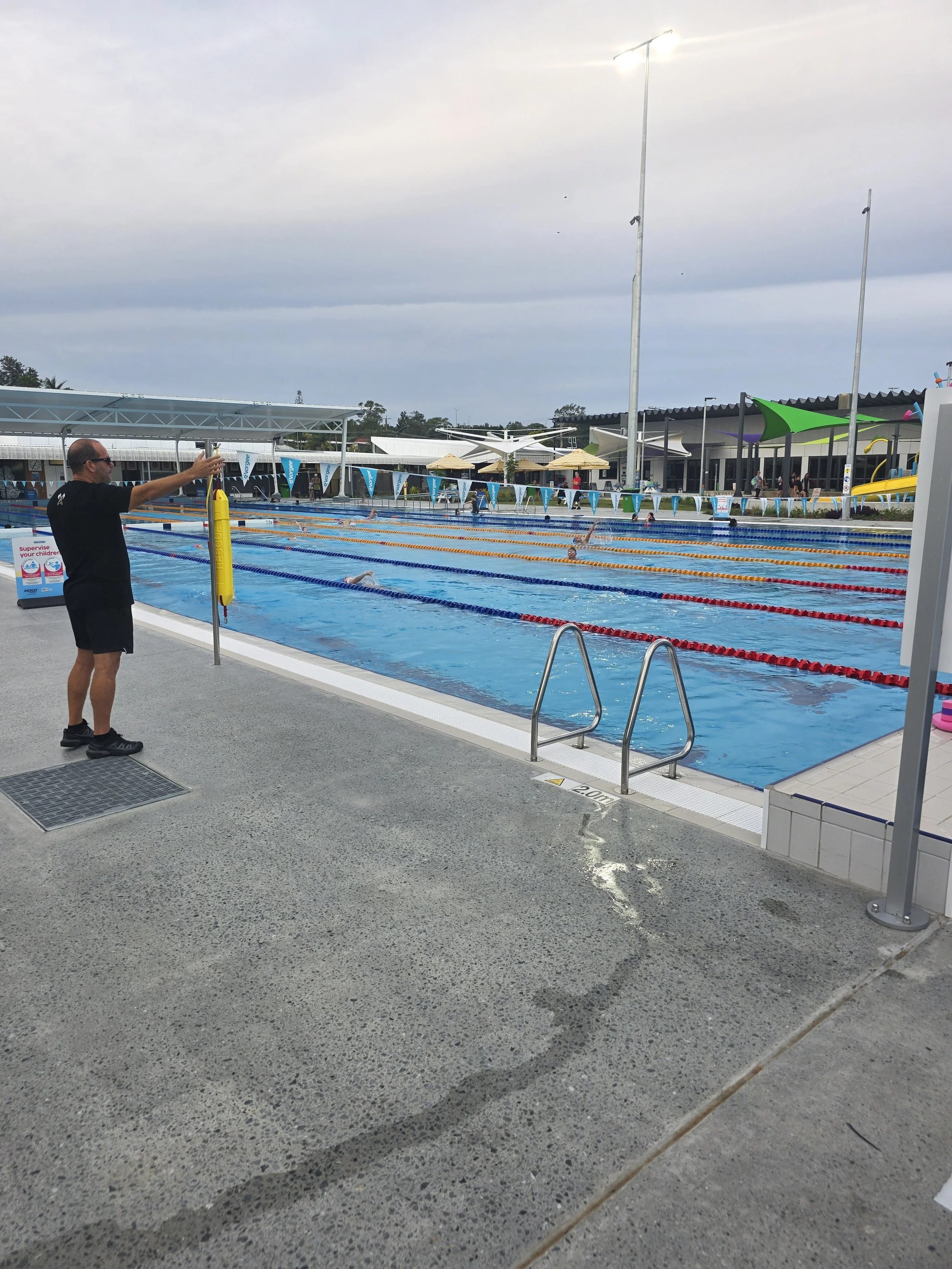 An outdoor swimming pool with swimmers, a lifeguard standing near the edge, and a poolside area with umbrellas and a playground. Overcast sky.