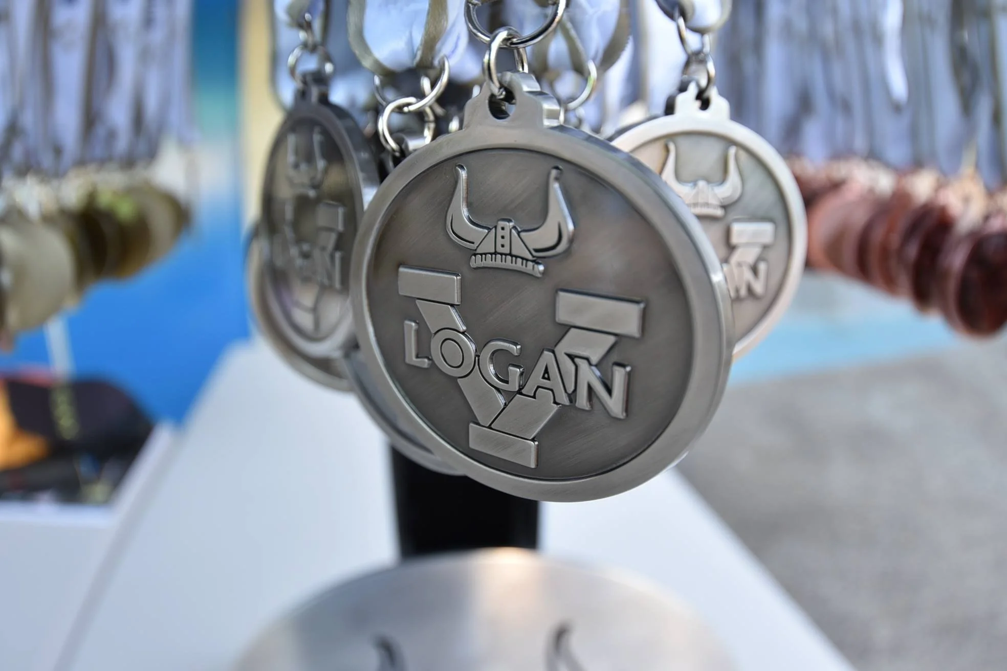 Close-up of a metal keychain with the name 'Logan' and a bull's head logo hanging on a display rack, with other keychains blurred in the background.