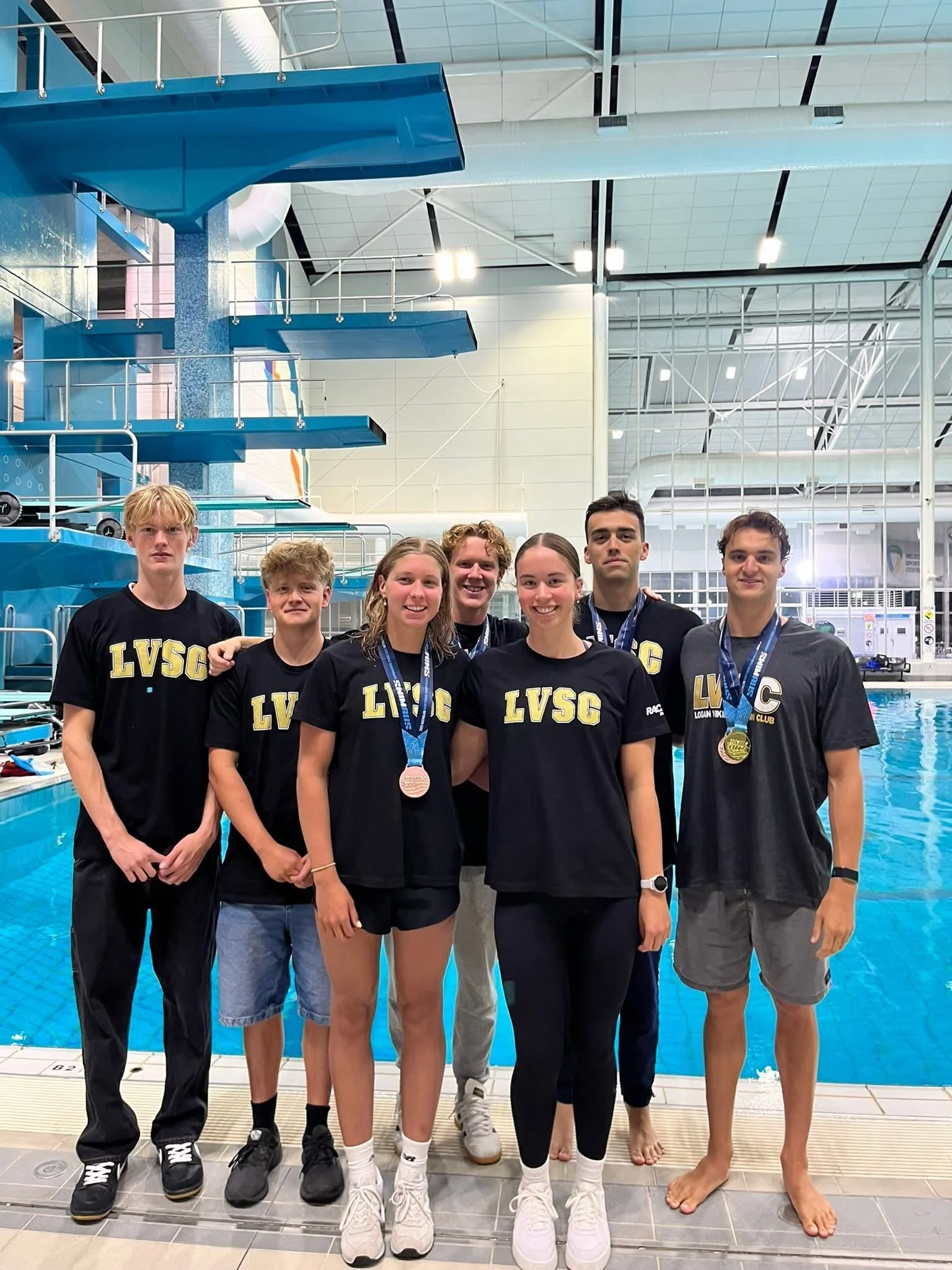 A group of seven young swimmers wearing black LVSC t-shirts and medals, standing on the pool deck of an indoor swimming pool.