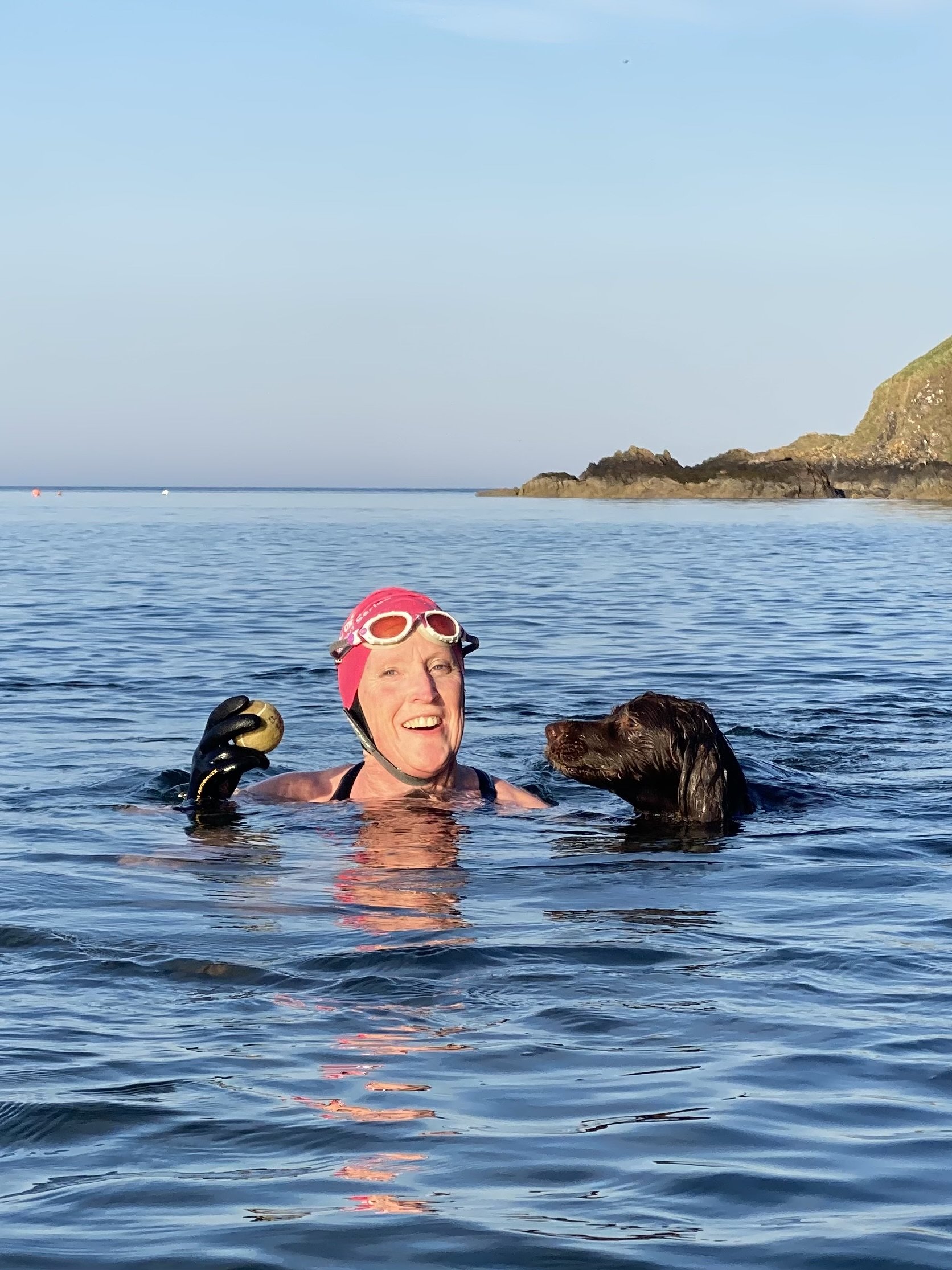 A woman wearing a pink swim cap and goggles is smiling while swimming in the ocean with a dog beside her. The background shows calm water, a rocky shoreline, and a clear sky.