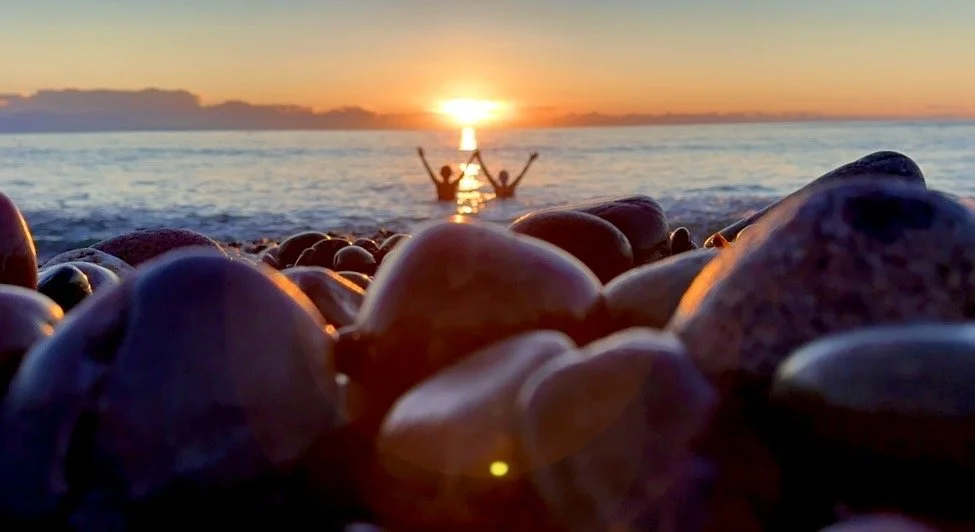 People enjoying a sunrise at the beach with their hands raised, seen from a low angle over colourful rocks in the foreground.