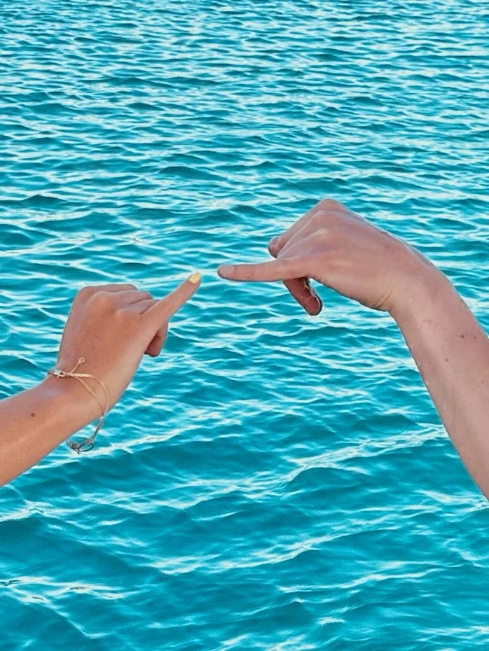 Two hands reaching over a blue ocean, playfully forming a heart shape.