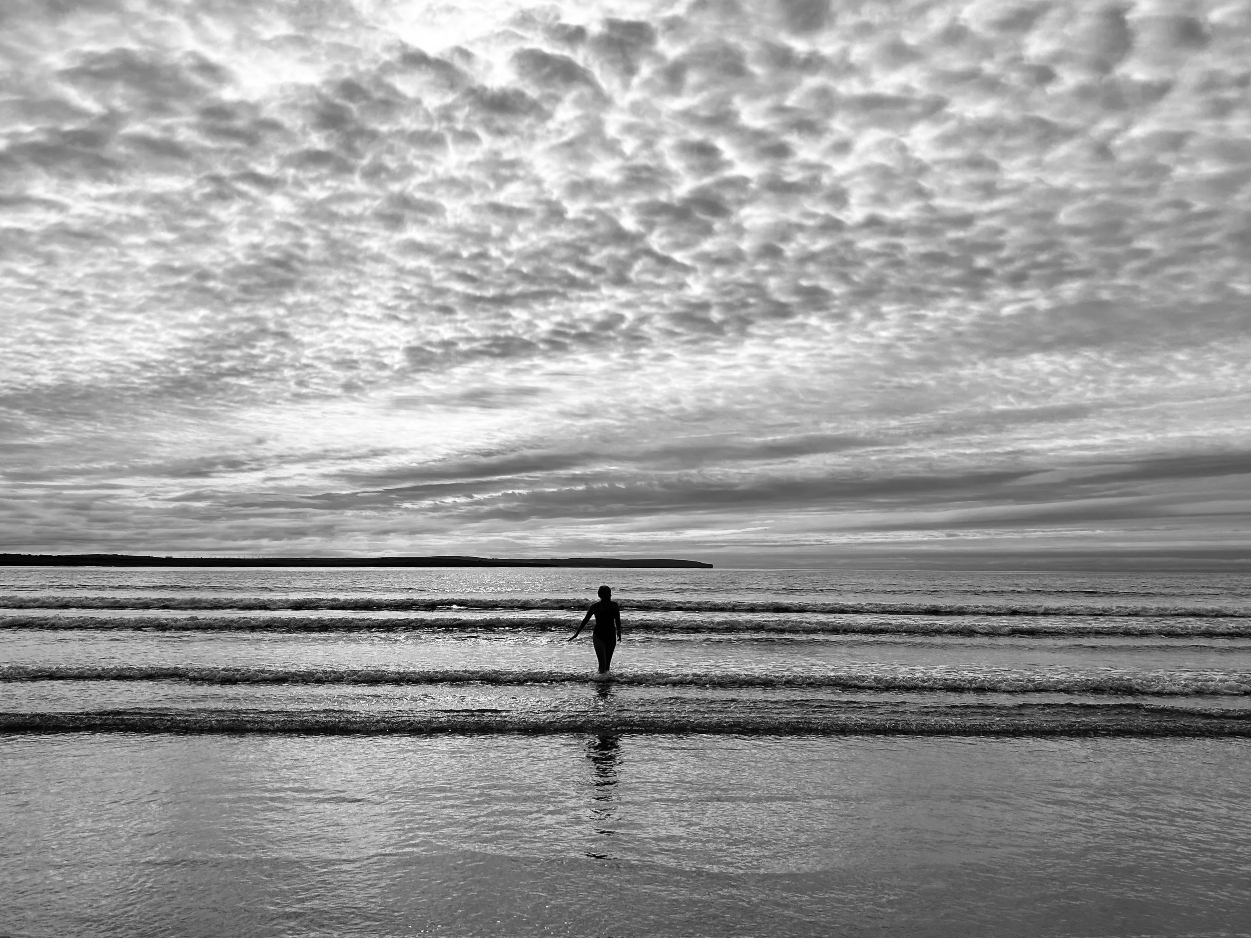 A person walking along the shoreline of a beach with a vast sky filled with textured clouds overhead.