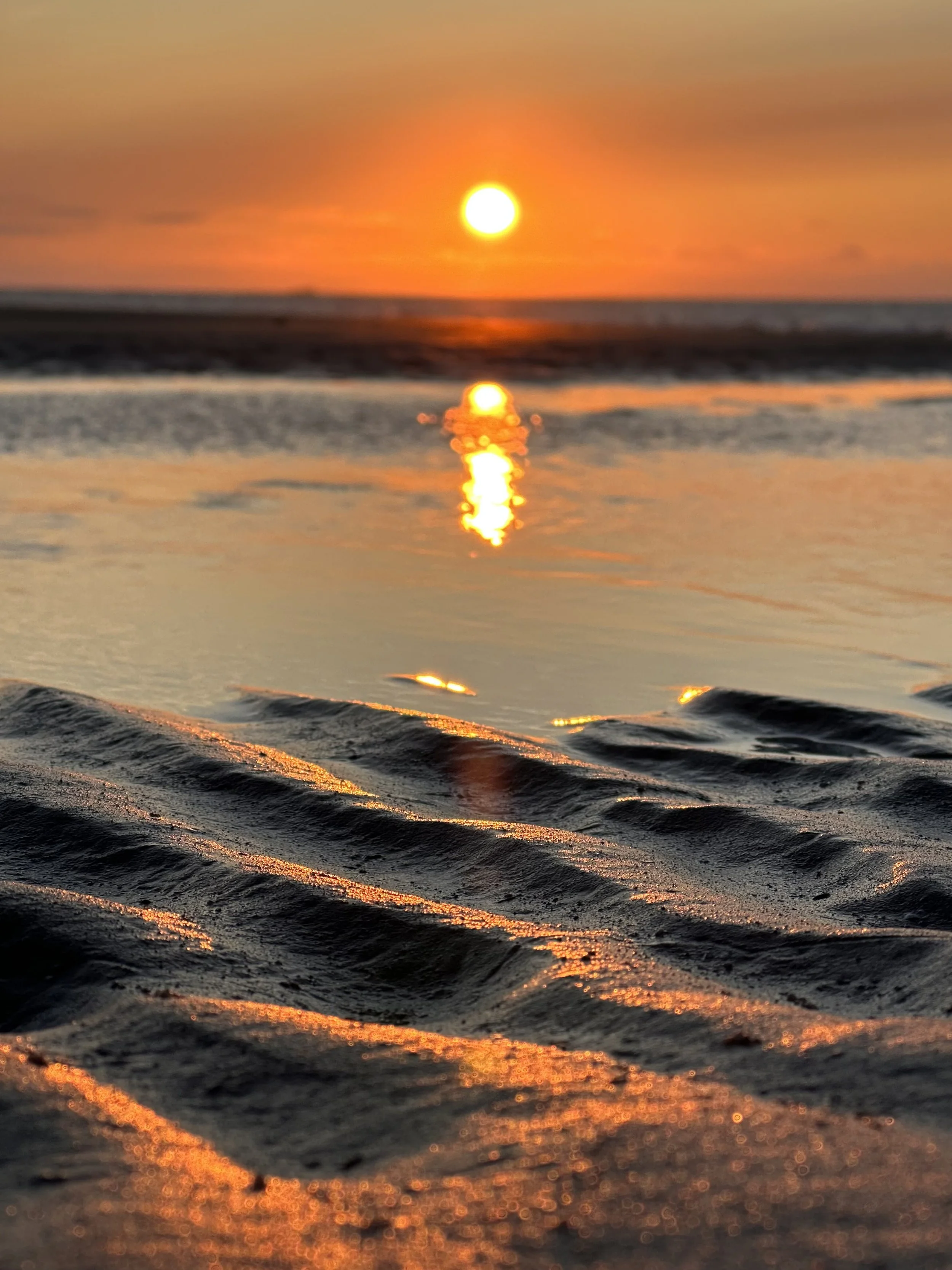 Sunset over the ocean with reflections on wet sand and gentle waves.