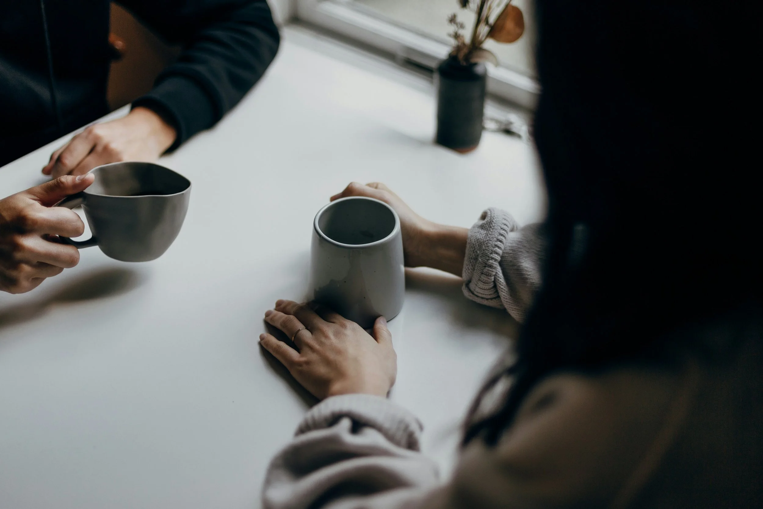 Two people exchanging cups at a white table, with a potted plant in the background.