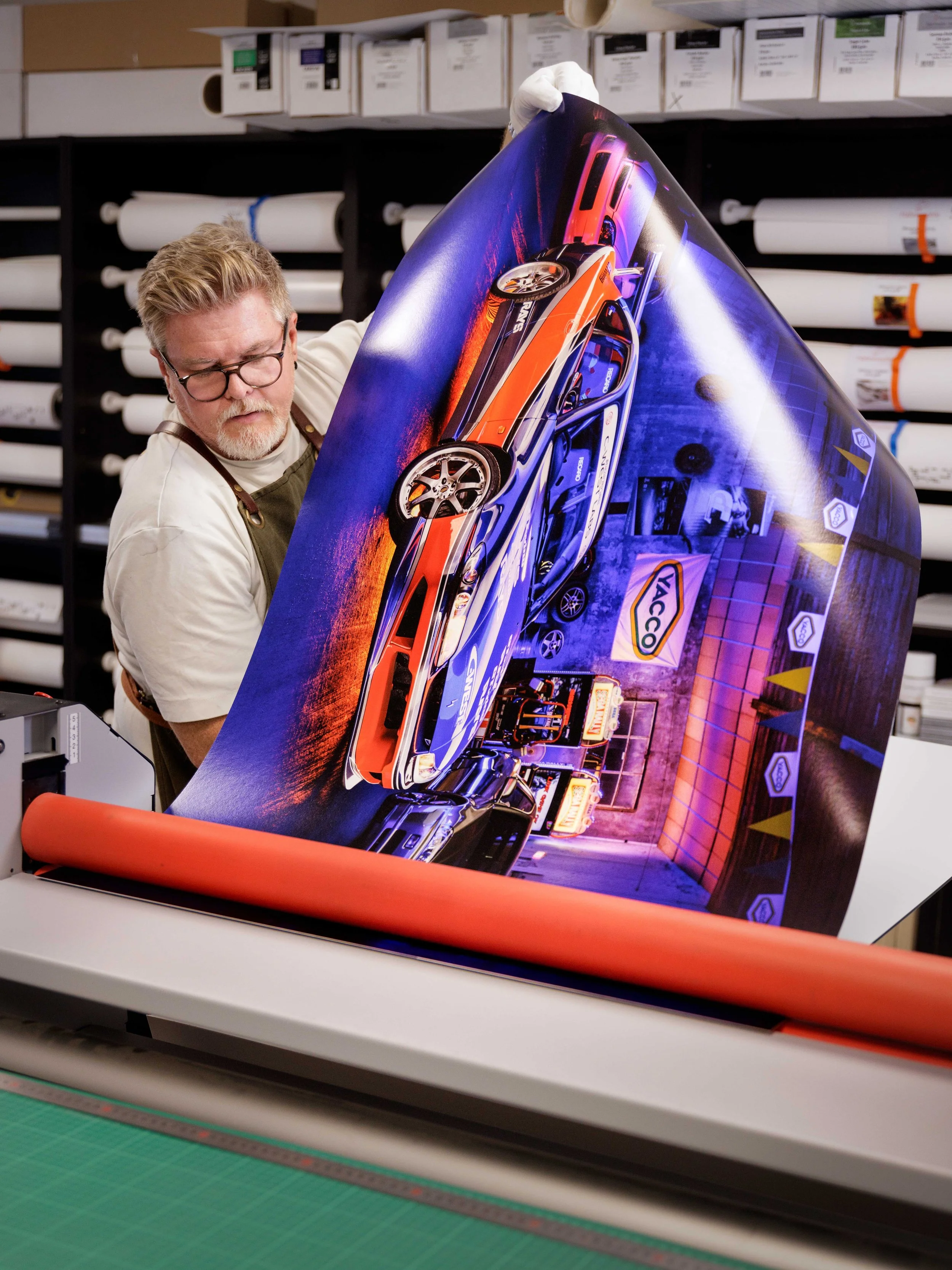 Un homme dans un atelier de production d'affiches suspend une grande affiche représentant une voiture de sport colorée, avec un fond urbain éclairé, dans une machine à imprimer.