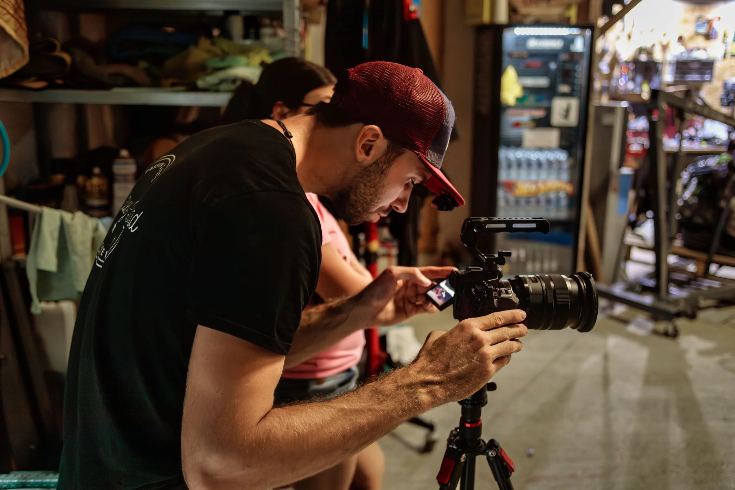 Un homme ajuste une caméra sur un trépied dans un atelier ou un lieu de travail, avec une femme en arrière-plan.