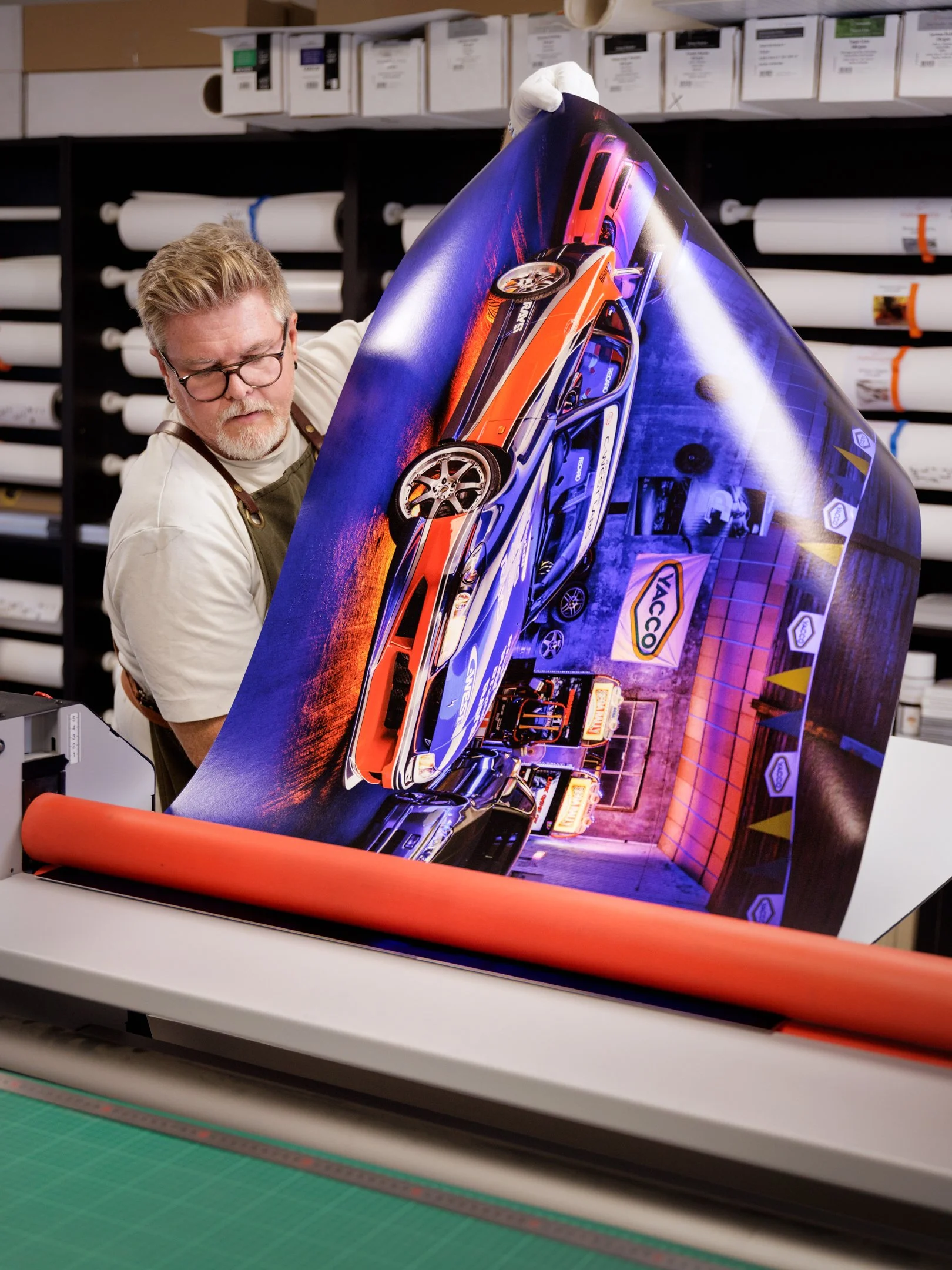 Une personne examine une affiche ou un poster grand format représentant une voiture de course, avec un fond urbain coloré et des logos, dans un environnement de studio ou d'atelier.