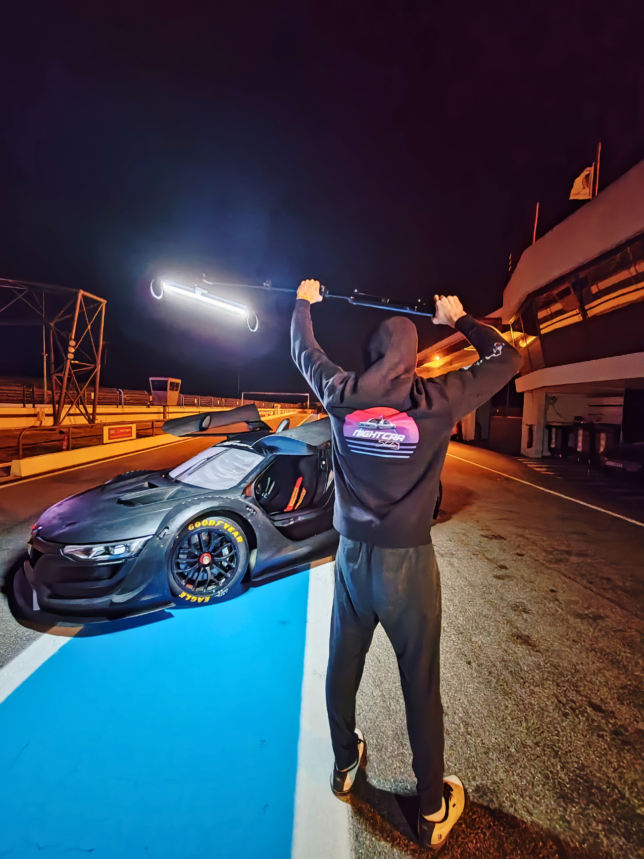 Une personne en hoodie noir avec un logo Nightcar sur le dos, debout sur la ligne de course d'un circuit, tenant un drapeau en l'air, derrière une maquette de voiture de course noire avec pneus Goodyear, sous un ciel nocturne.
