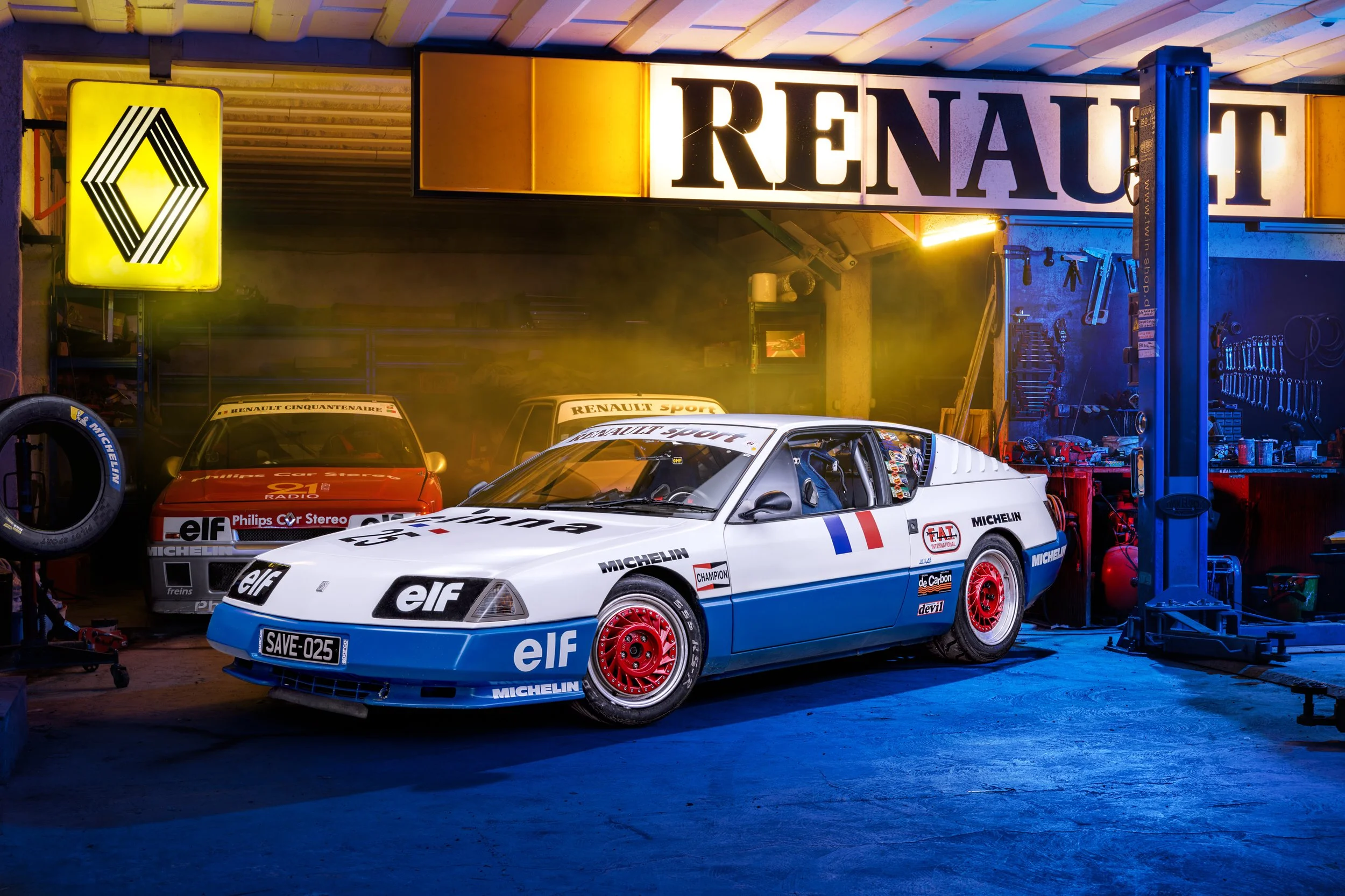 Une voiture de course emblématique dans un garage, avec deux autres voitures derrière. Le garage est équipé d'outils et d'équipements de mécanique, et une grande enseigne Renault est visible en arrière-plan.
