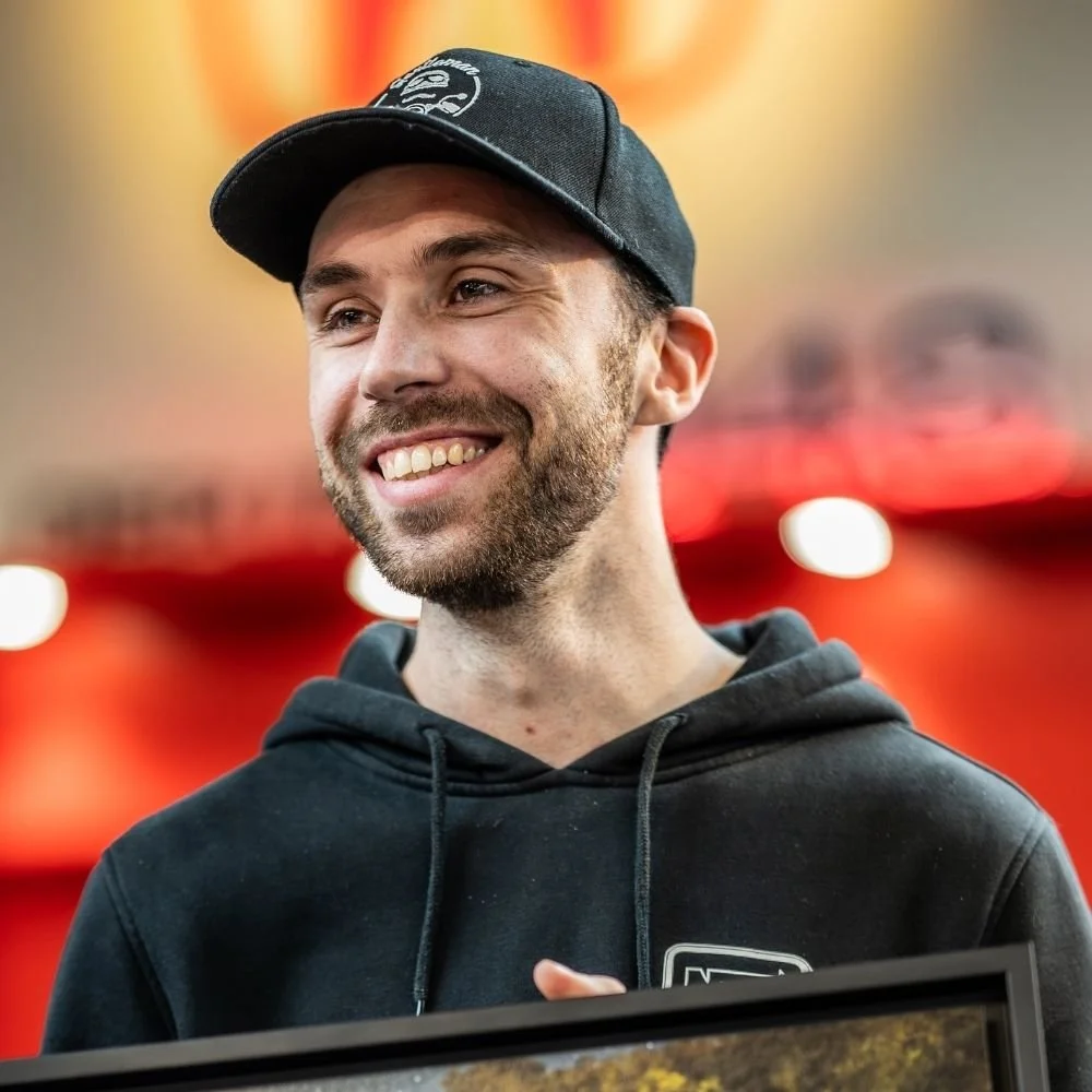 Un homme souriant porte une casquette noire et un sweat à capuche noir, avec un fond rouge flou.