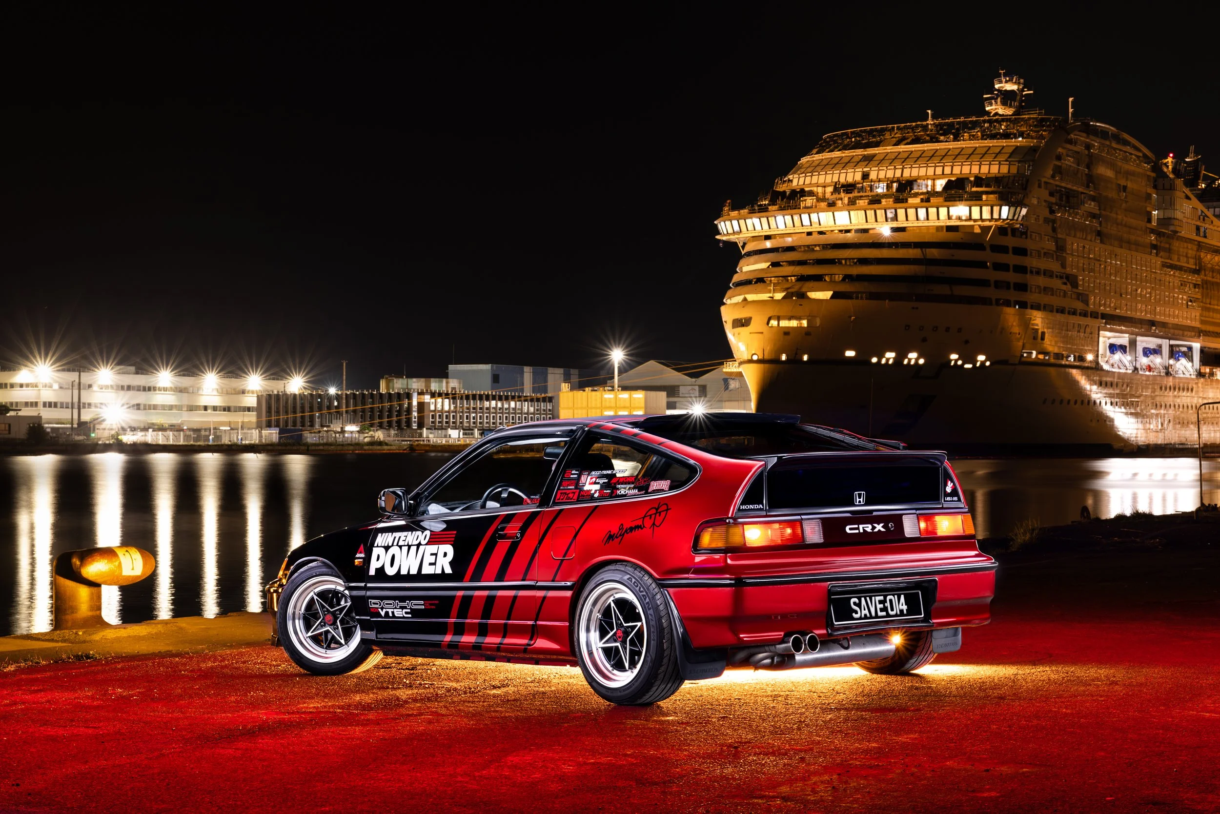 Voiture de sport rouge et noire modifiée stationnée près du quai la nuit, avec un grand navire de croisière illuminé en arrière-plan au port.
