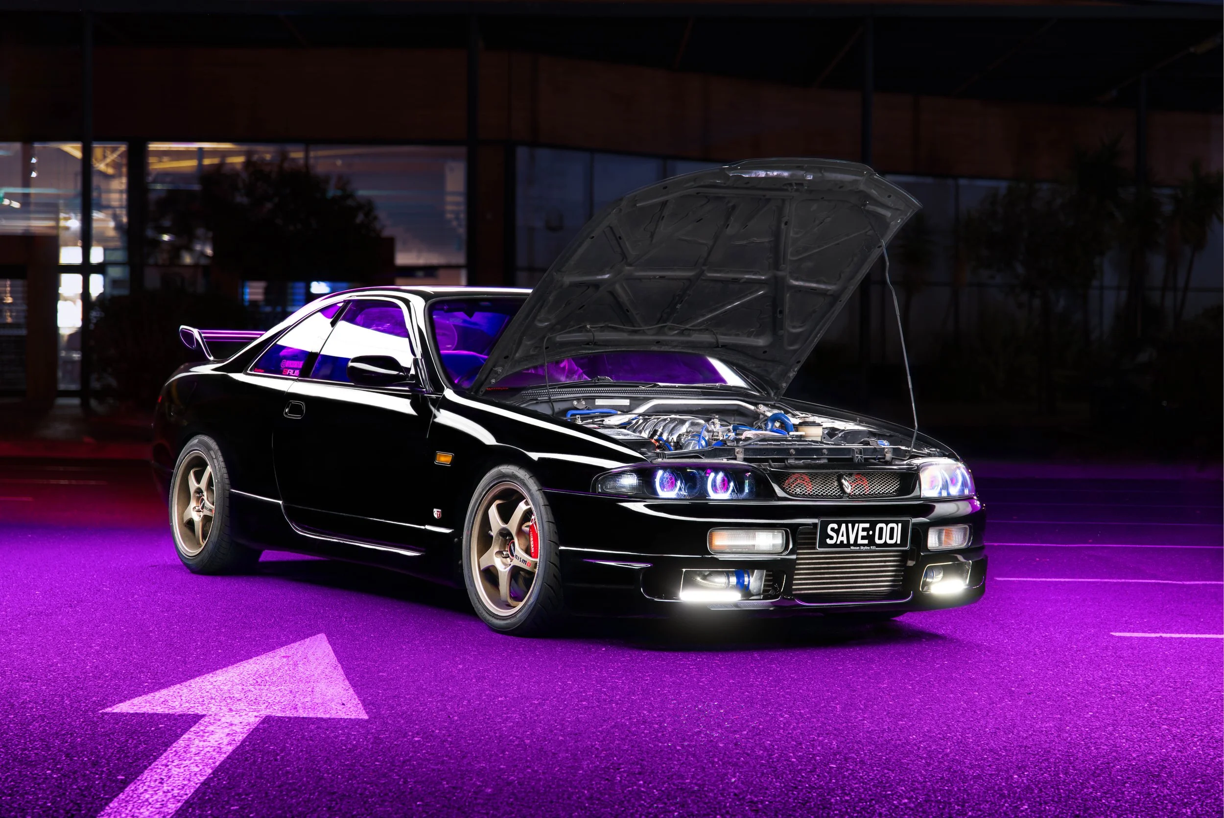 Voiture noire modifiée avec capot relevé, illuminée par des lumières violettes, stationnée dans une zone urbaine la nuit, sur une route avec une flèche blanche pointant vers la gauche.