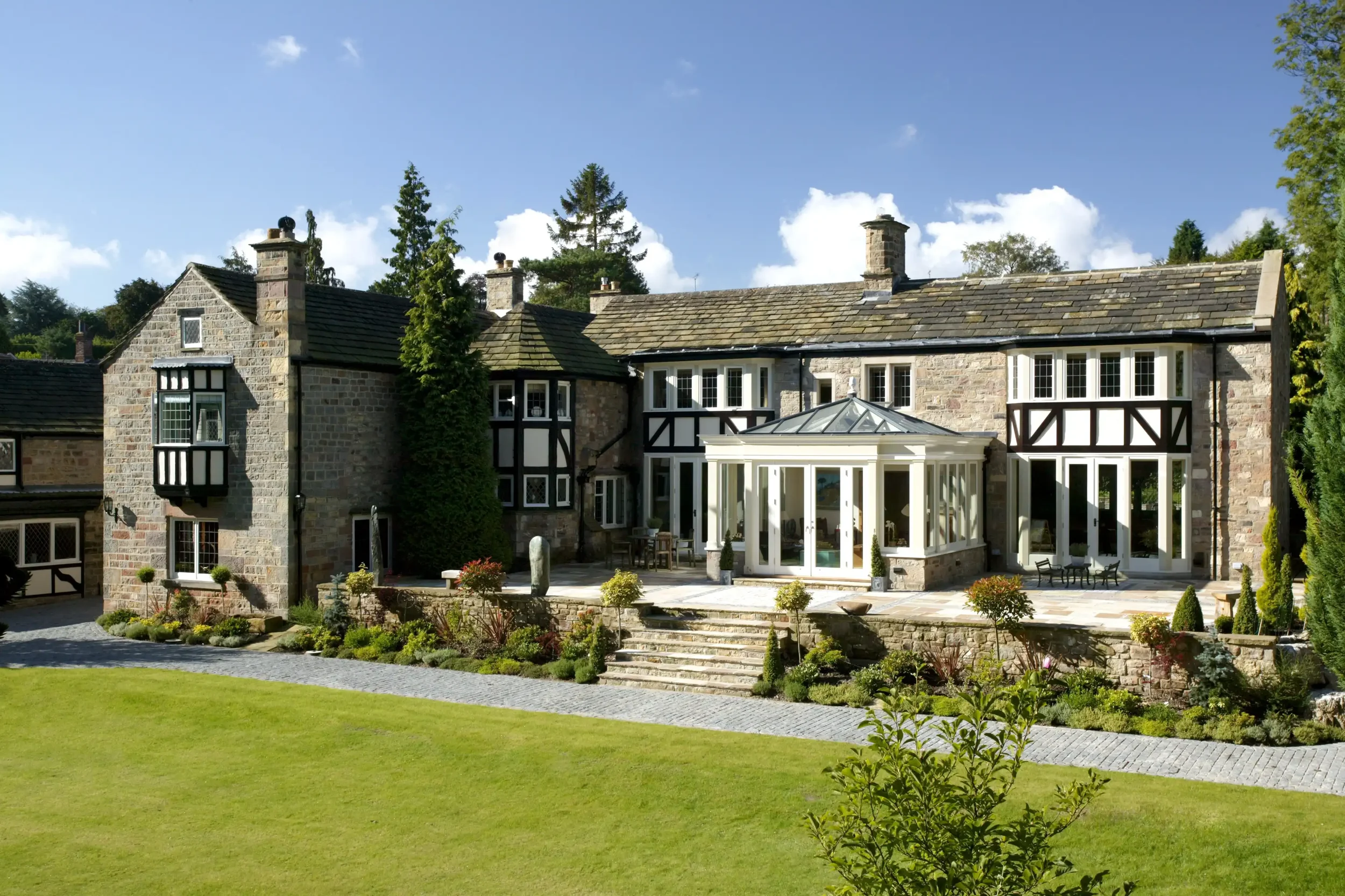Classical painted timber orangery with a roof lantern viewed from the garden of a large stone and timber-detailed house.