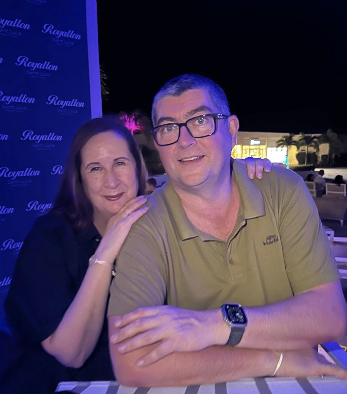 A man and a woman posing together outdoors at night, with a backdrop that says "Royallon" and some buildings and palm trees in the background.