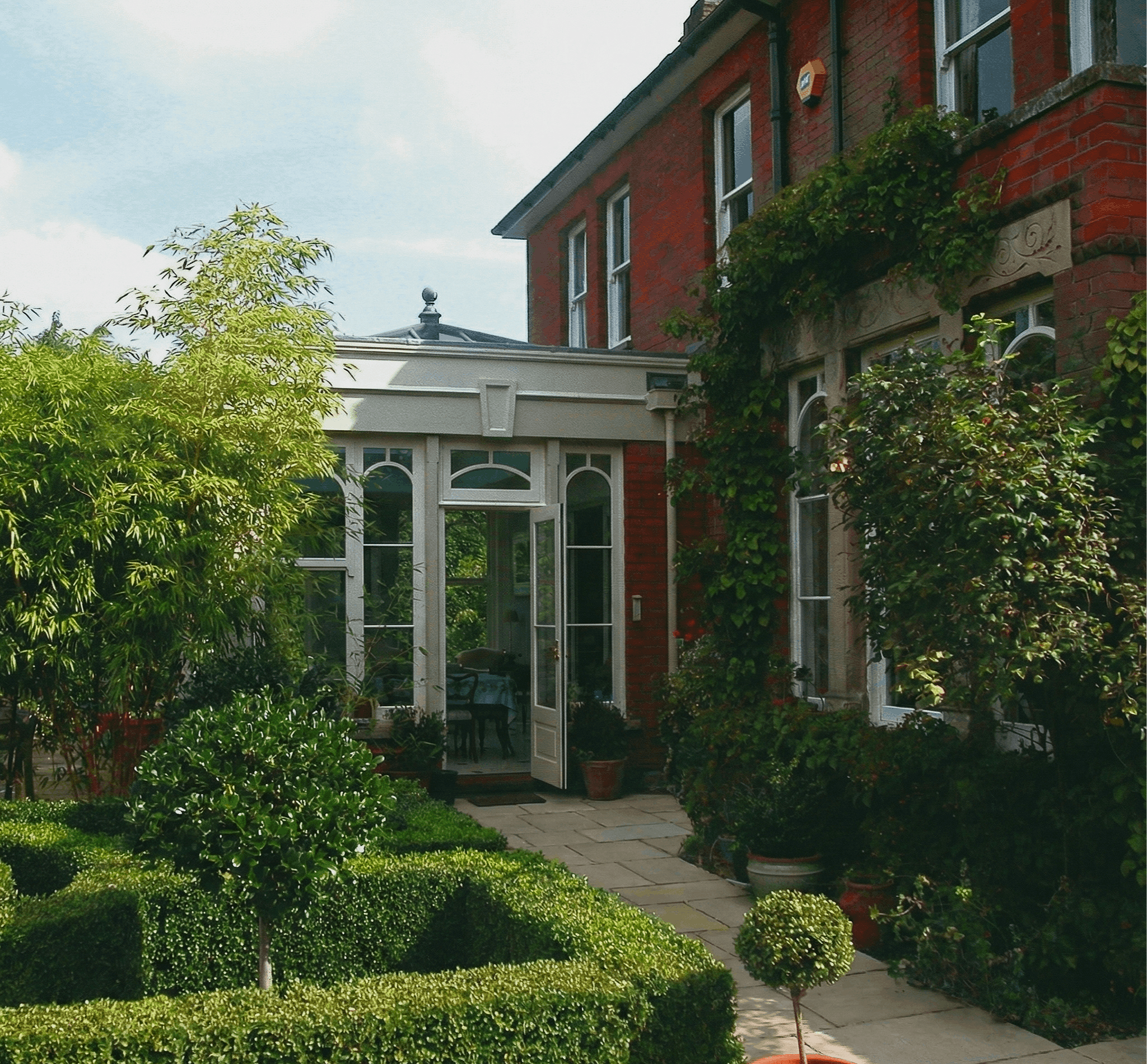 Red brick house with garden, open patio door, and greenery including trees, bushes, and potted plants.