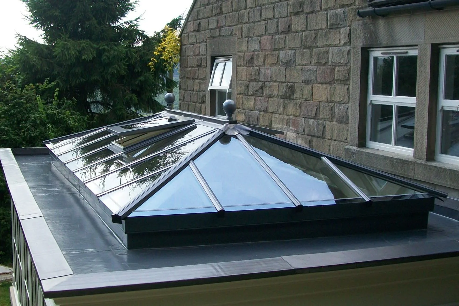 A glass and metal skylight on a flat roof of a brick building, with multiple glass panels and decorative black fixtures on top, reflecting the sky and nearby trees.