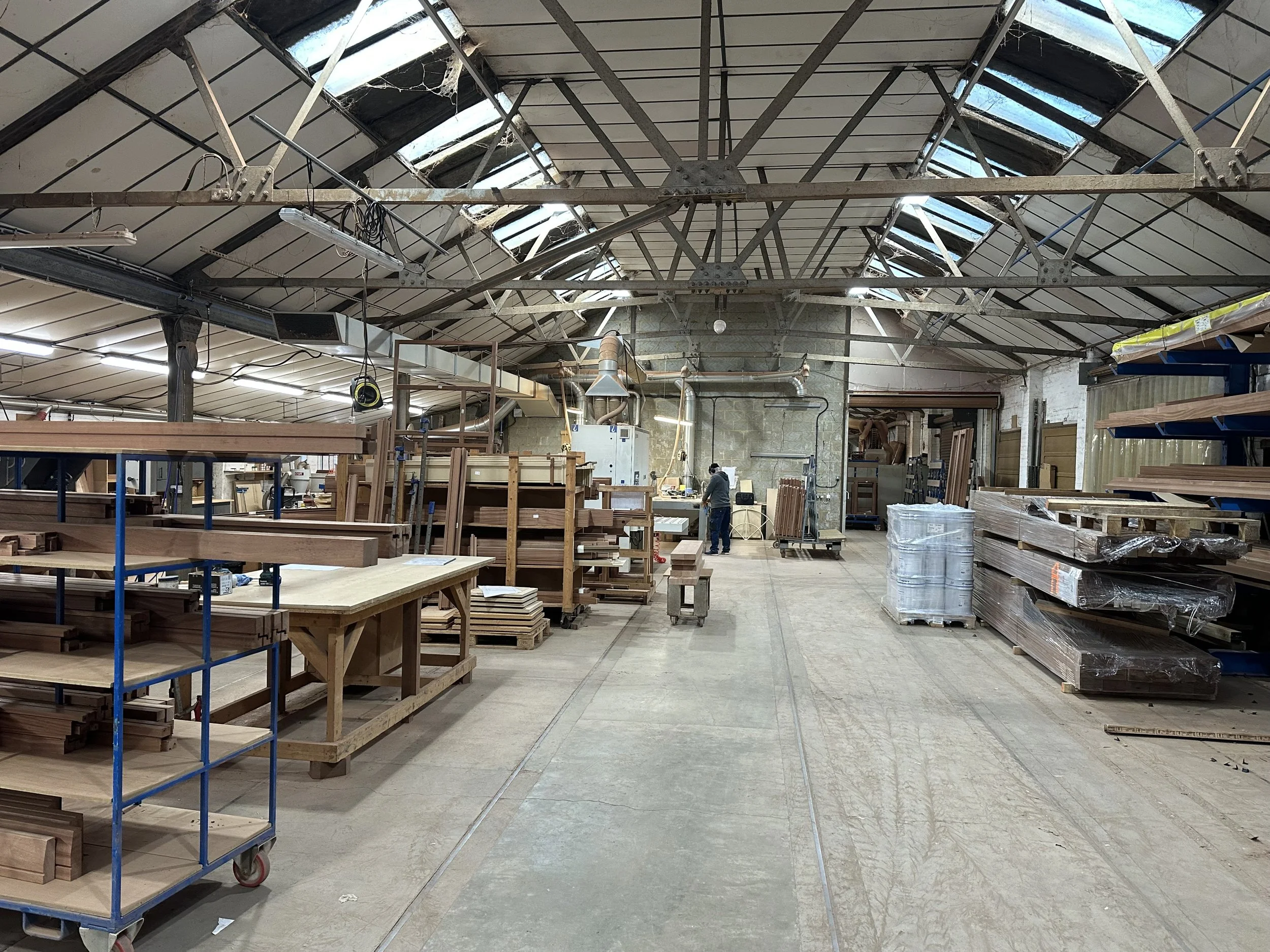 Inside a woodworking shop with various shelves, stacks of wood, and a worker at a workbench under a high, metal-framed roof with skylights.