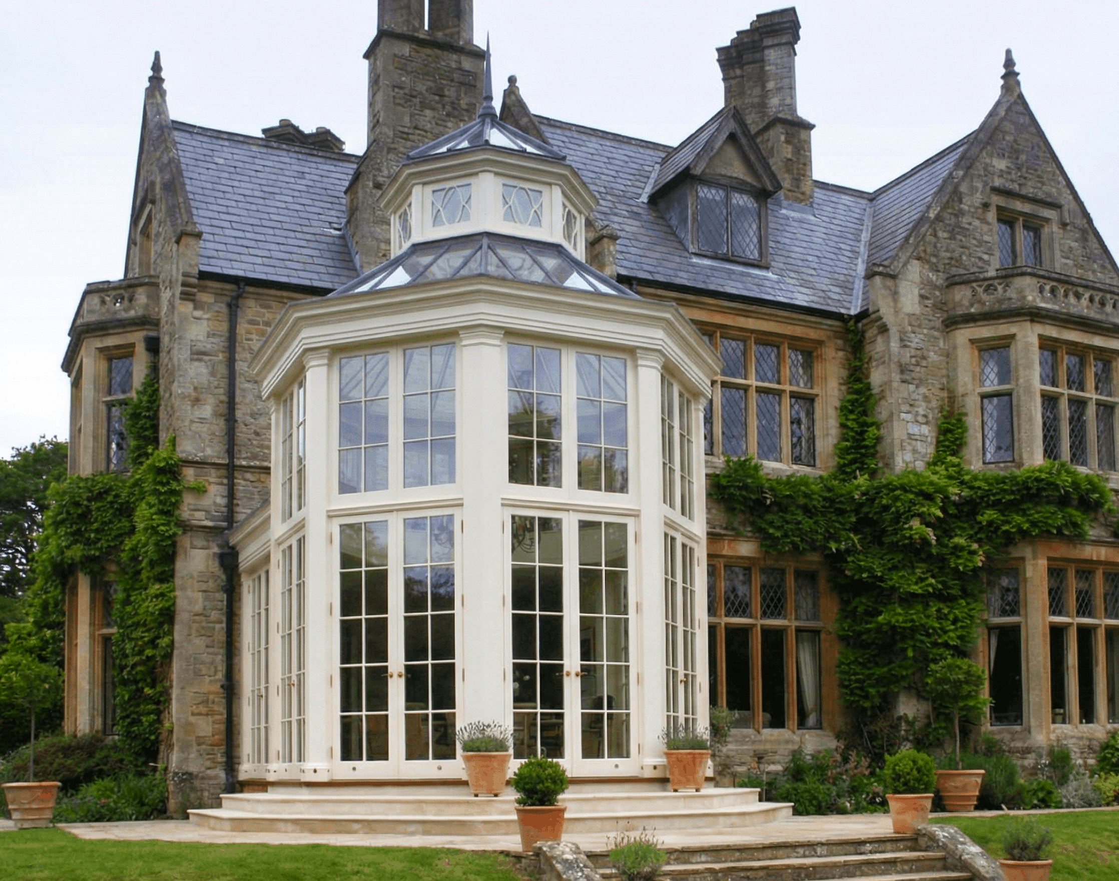 A large, historic stone mansion with multiple chimneys and a prominent white glass conservatory in the front. The house features gothic-style architecture with pointed turrets and leaded glass windows partly covered with green climbing vines. There are outdoor potted plants and a well-maintained lawn in front of the house.