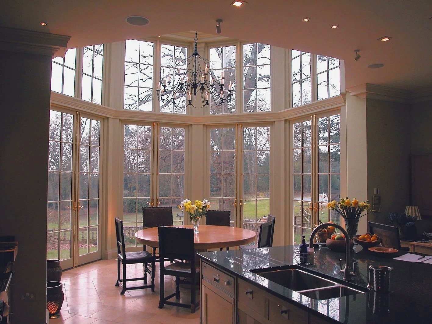 Bright kitchen with large bay window and dining area, featuring a chandelier, table with flowers, and view of trees outside.