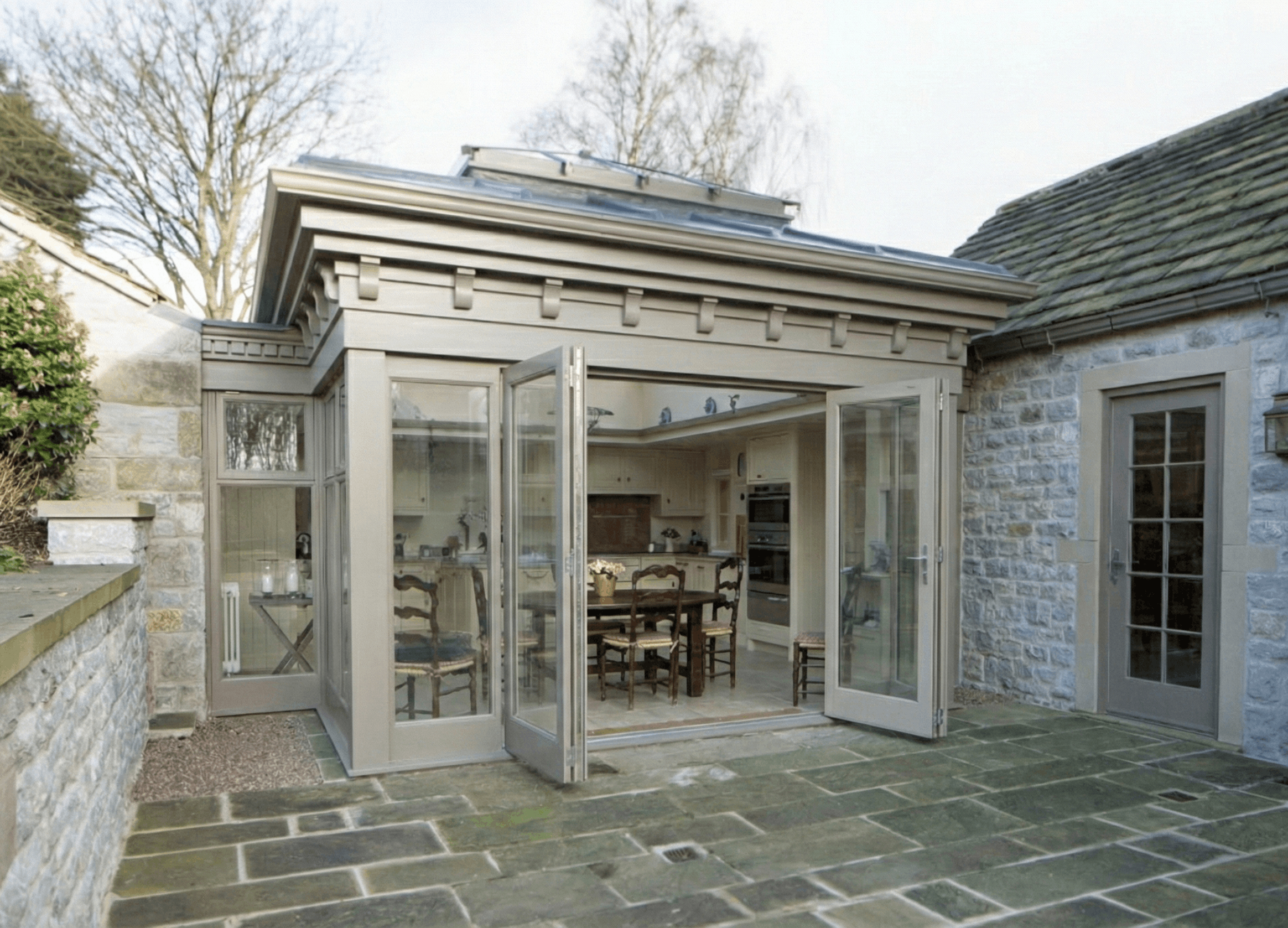 Painted hardwood kitchen extension with wide glazed doors opening onto a stone courtyard beside a period house.