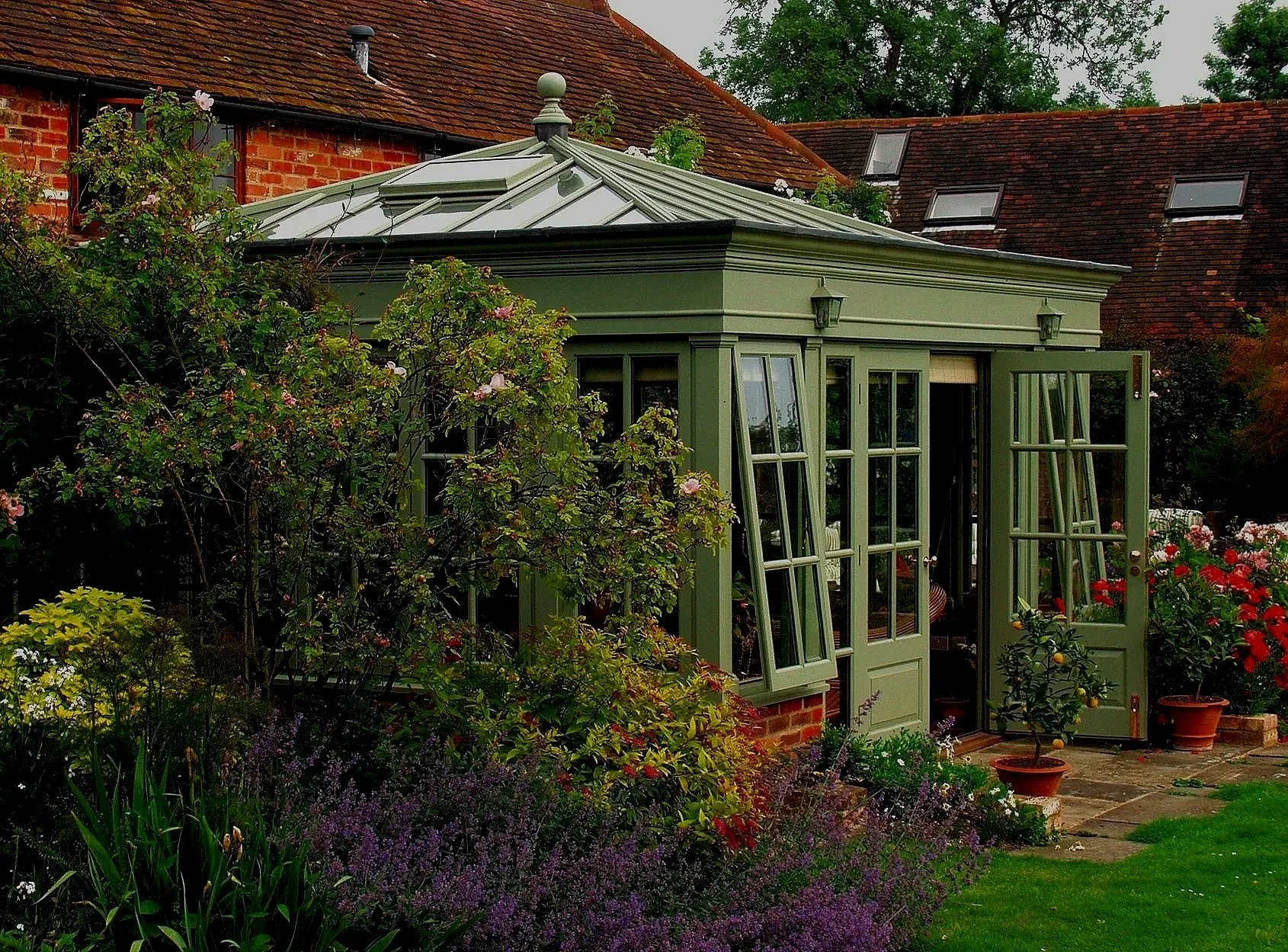 Green-painted hardwood orangery with roof lantern and French doors surrounded by a mature cottage garden.
