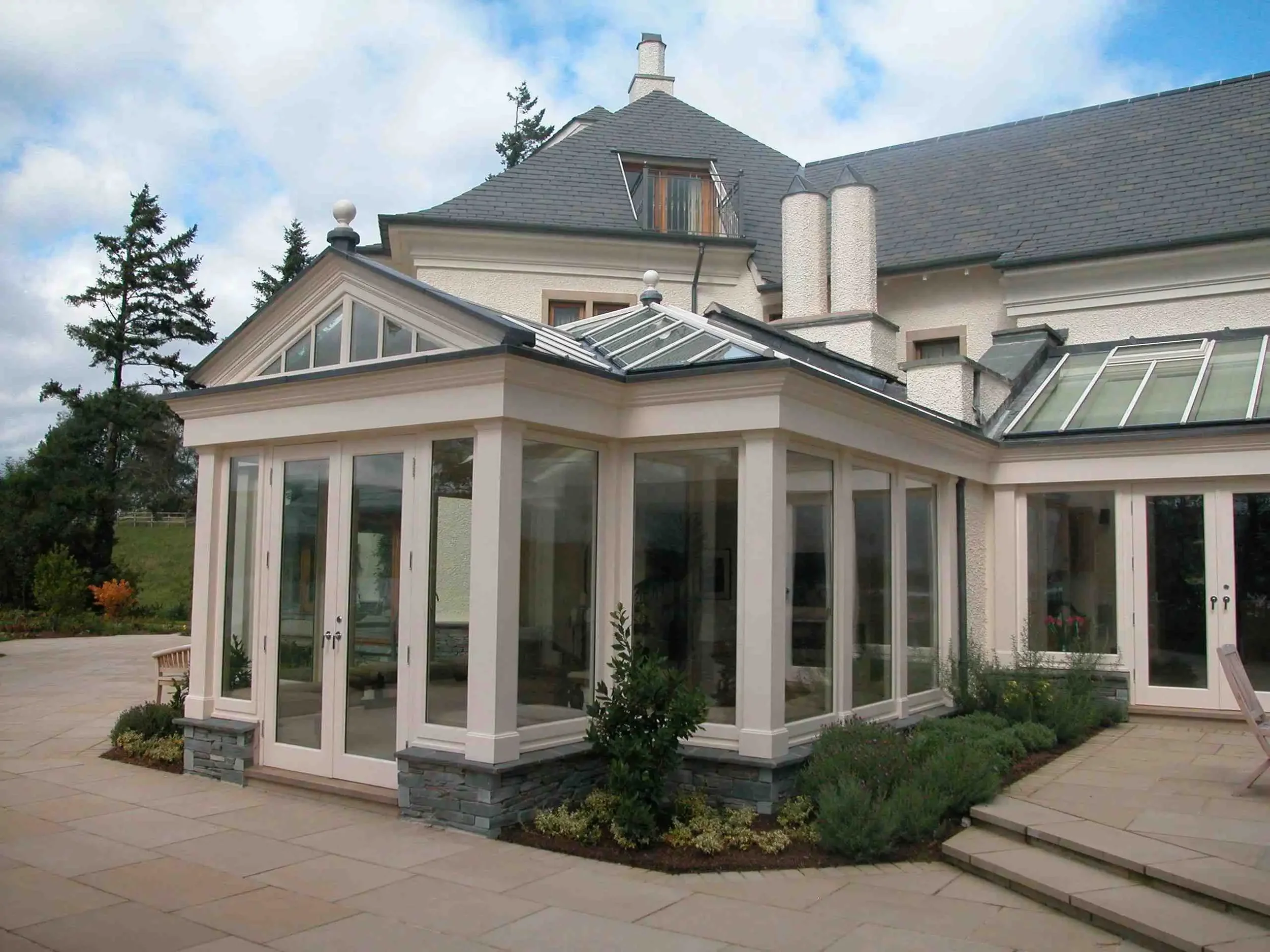 Large pale-painted hardwood glazed extension with roof lanterns attached to a substantial rendered country home.
