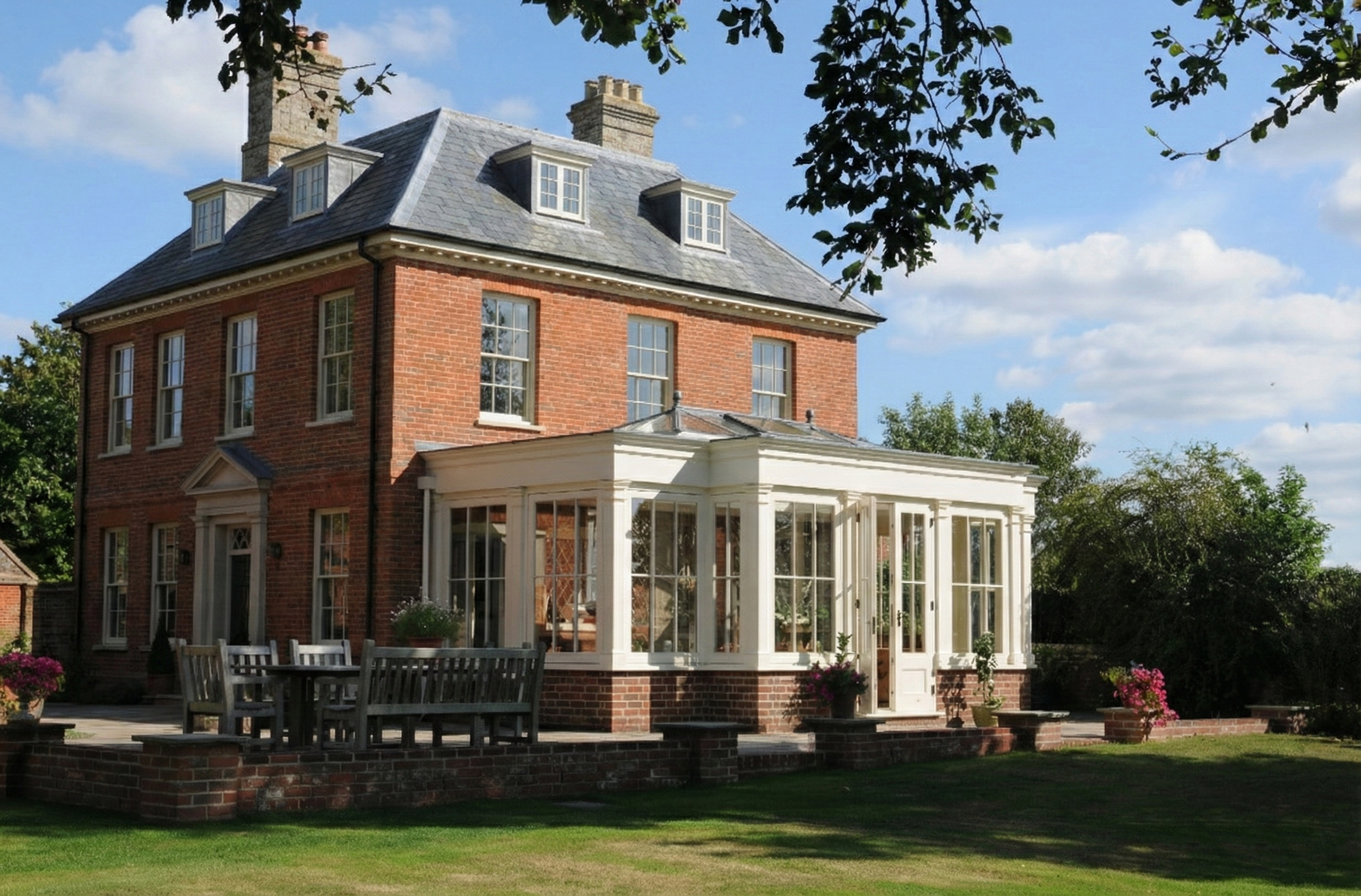 A traditional red brick house with multiple windows, a slate roof with dormer windows, and a white sunroom or porch extension with large windows, surrounded by a well-maintained lawn and some potted plants, under a partly cloudy sky.