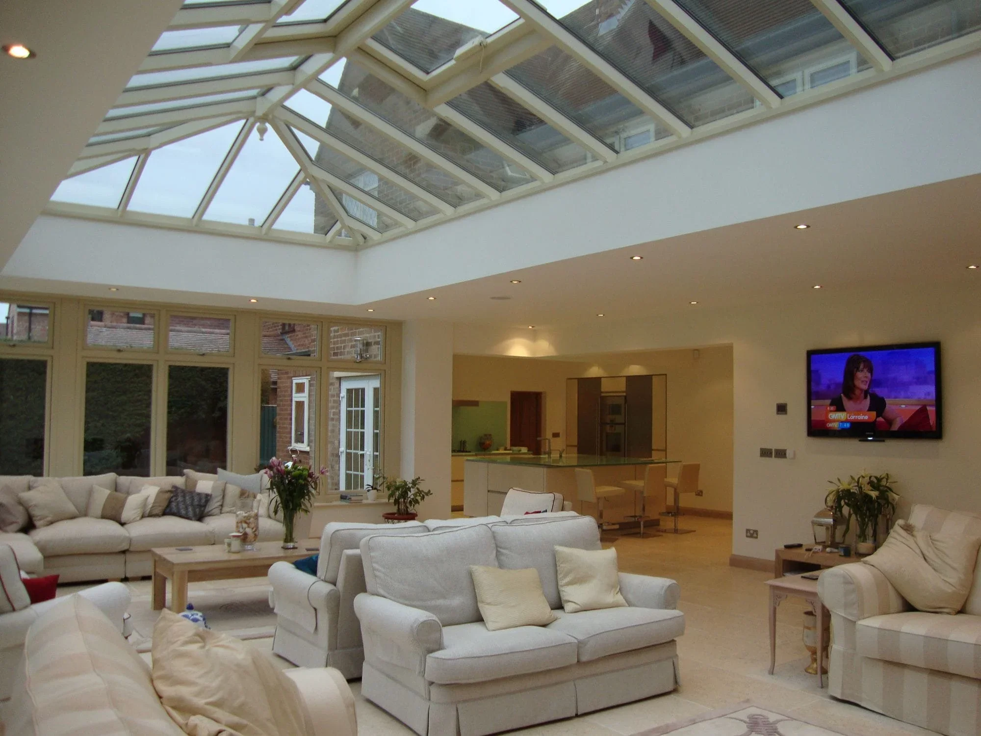 Large open-plan family living space beneath an expansive glazed roof with seating area, dining space and kitchen beyond.