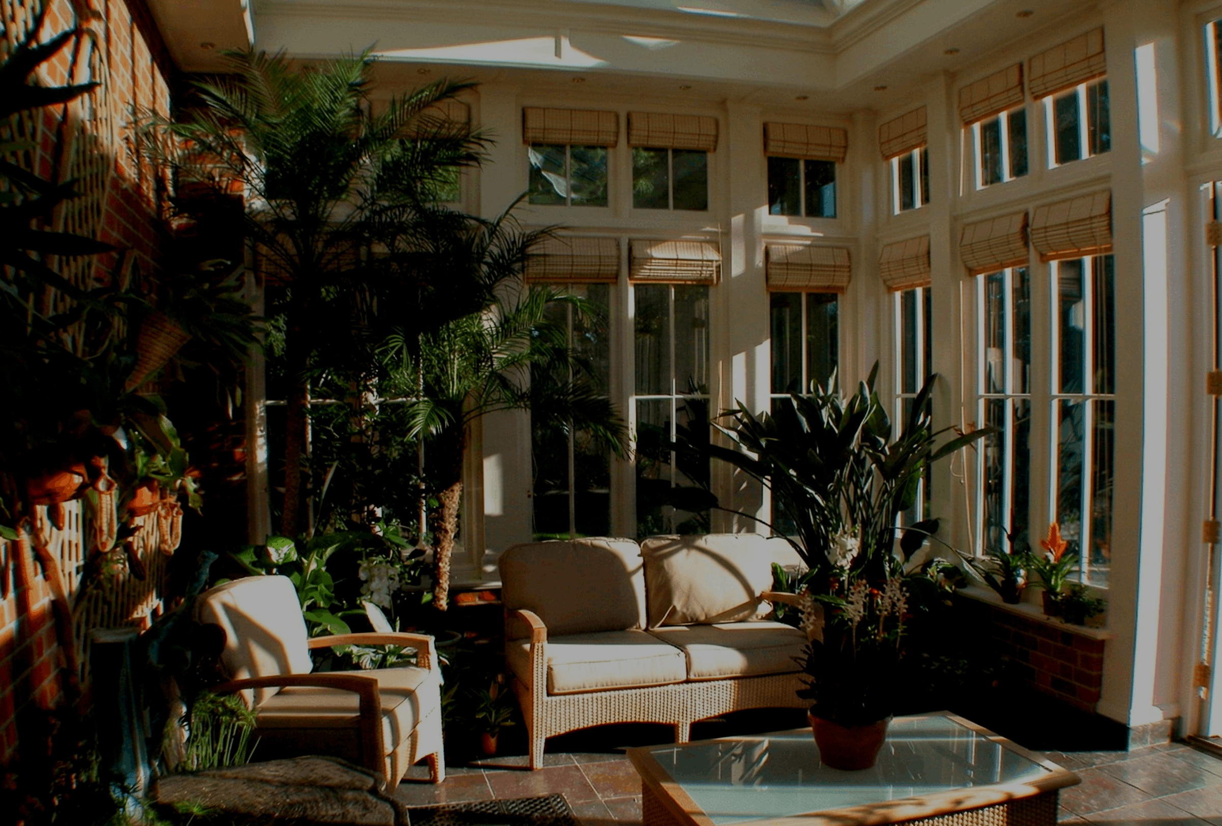 Sunlit sunroom with large windows, a glass-top coffee table, potted plants, and wicker furniture, including a loveseat and an armchair.