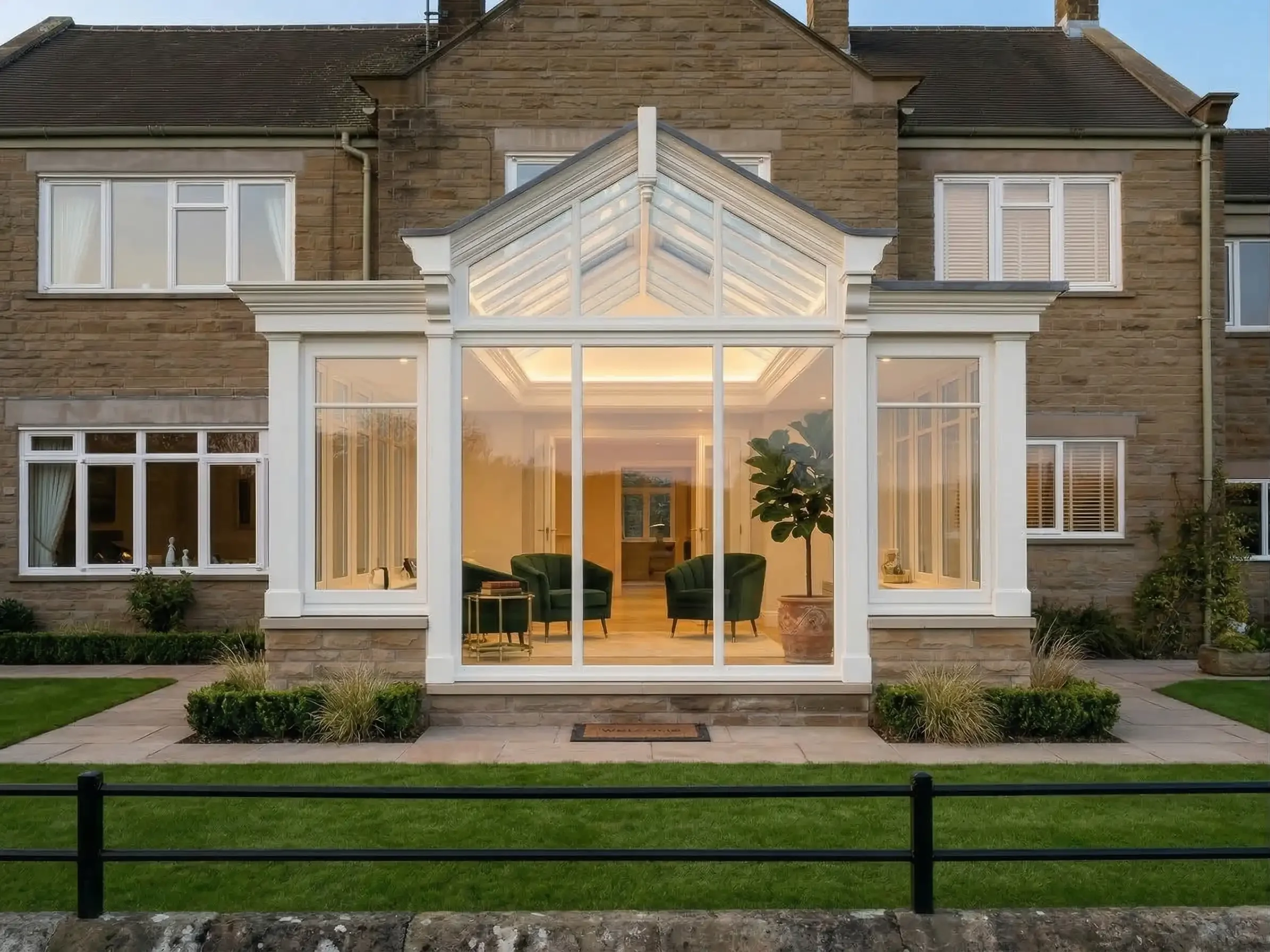 A white timber gable-fronted garden room extension built on a matching stone dwarf wall next to an old stone house.