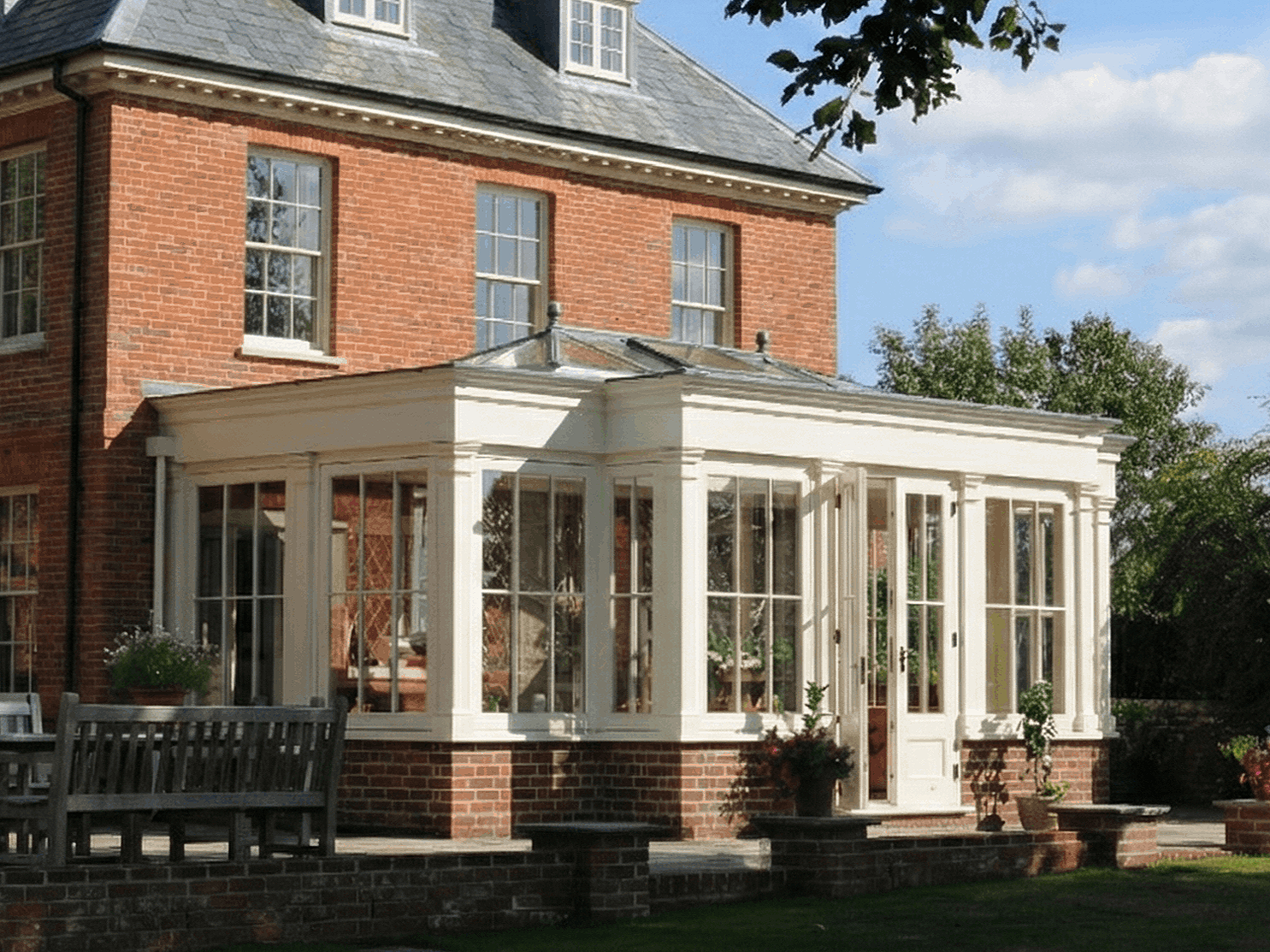 A brick house with a white enclosed patio or sunroom with large windows, outdoor plants, and a wooden bench in the garden, under a partly cloudy sky.