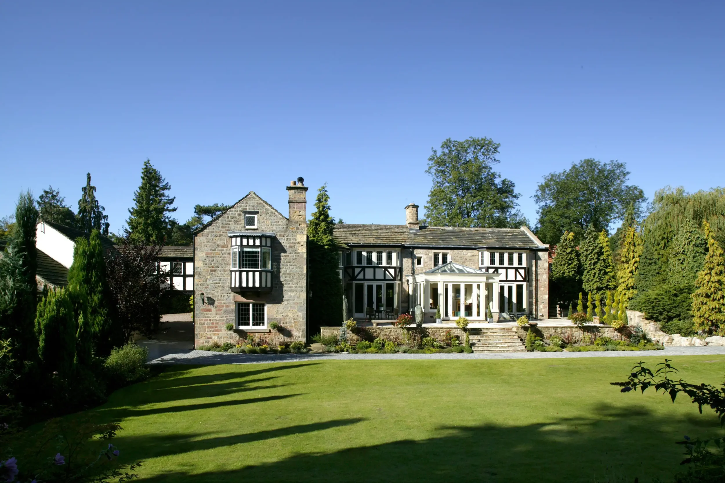 Wide garden view of a large stone manor house with a classical painted timber orangery centred on the rear terrace.