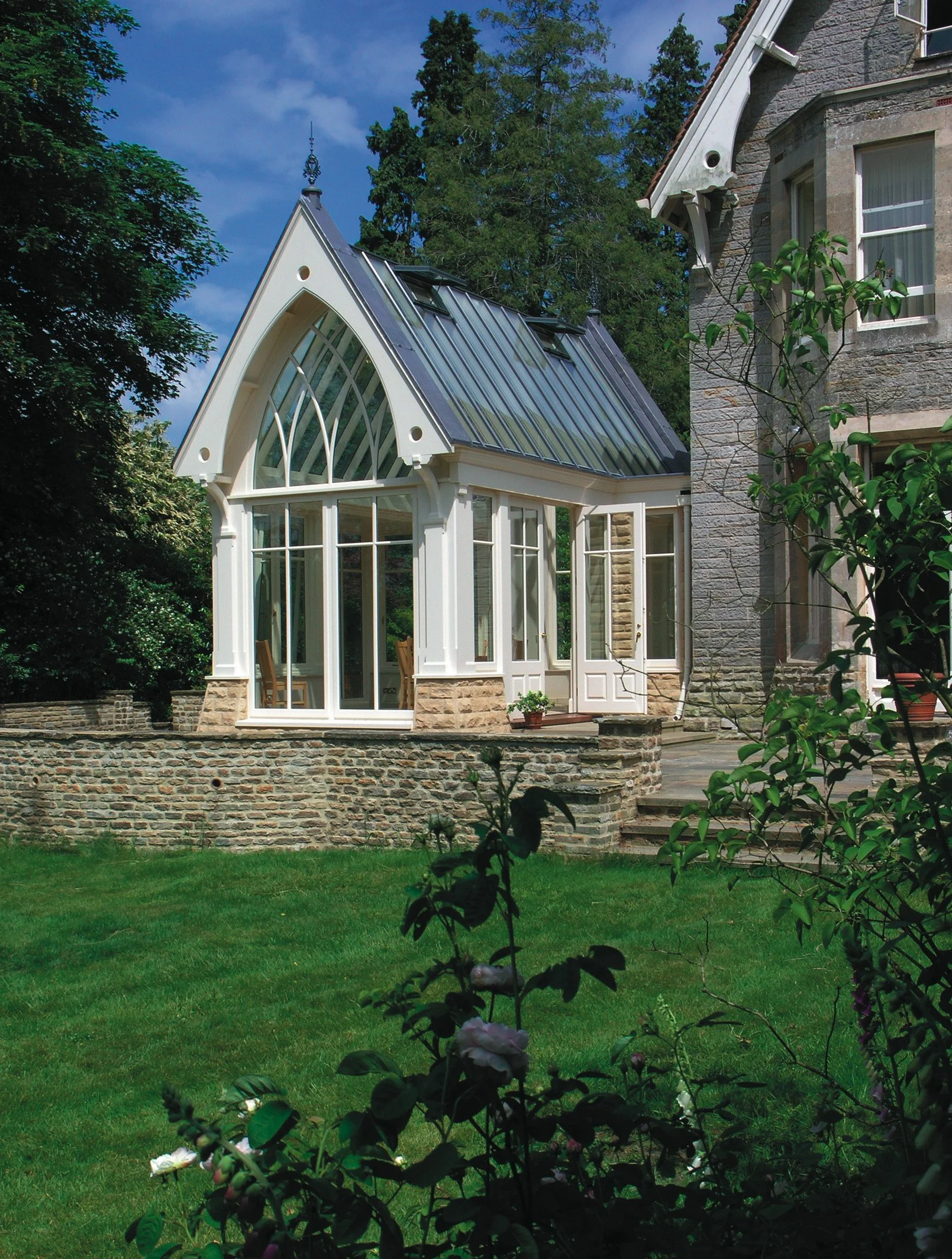 A house with a distinctive glass-enclosed sunroom or conservatory attached, featuring large windows and a steep metal roof, surrounded by a lush green lawn and trees.