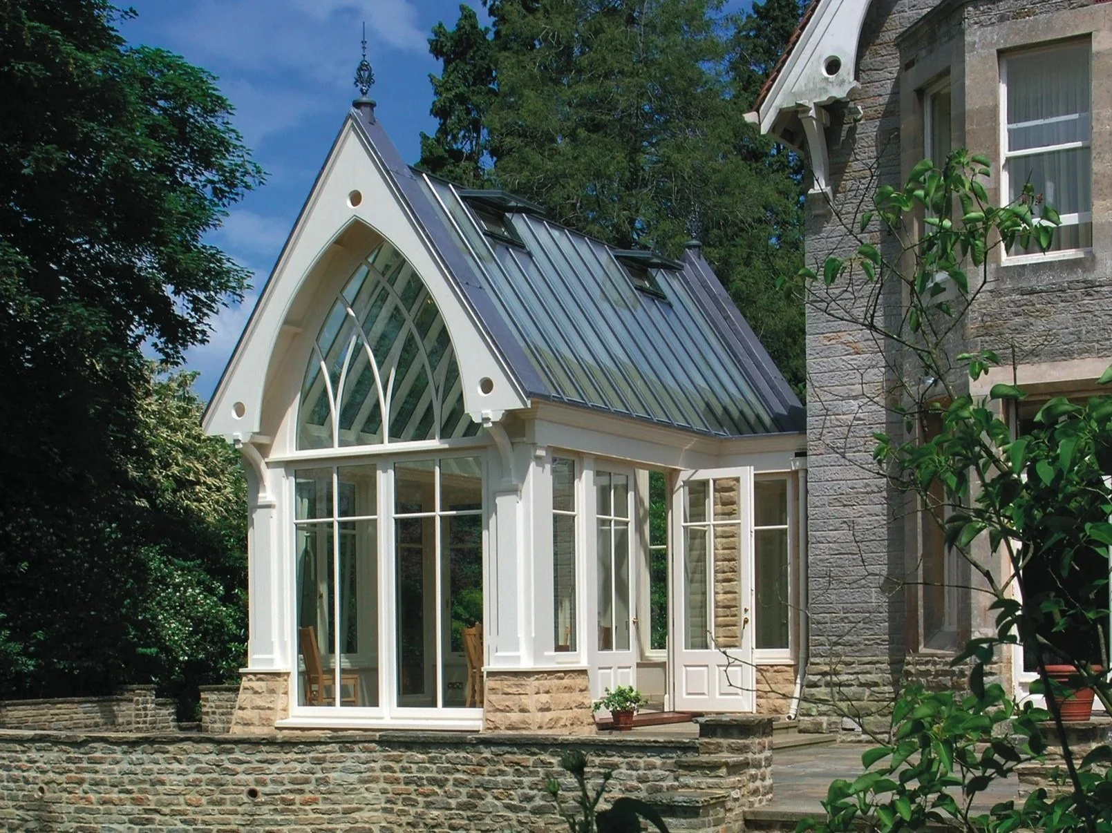 A house with a tall, pointed roof with metal panels, a large arched window, white framed glass walls, and stone foundation, surrounded by greenery and trees.