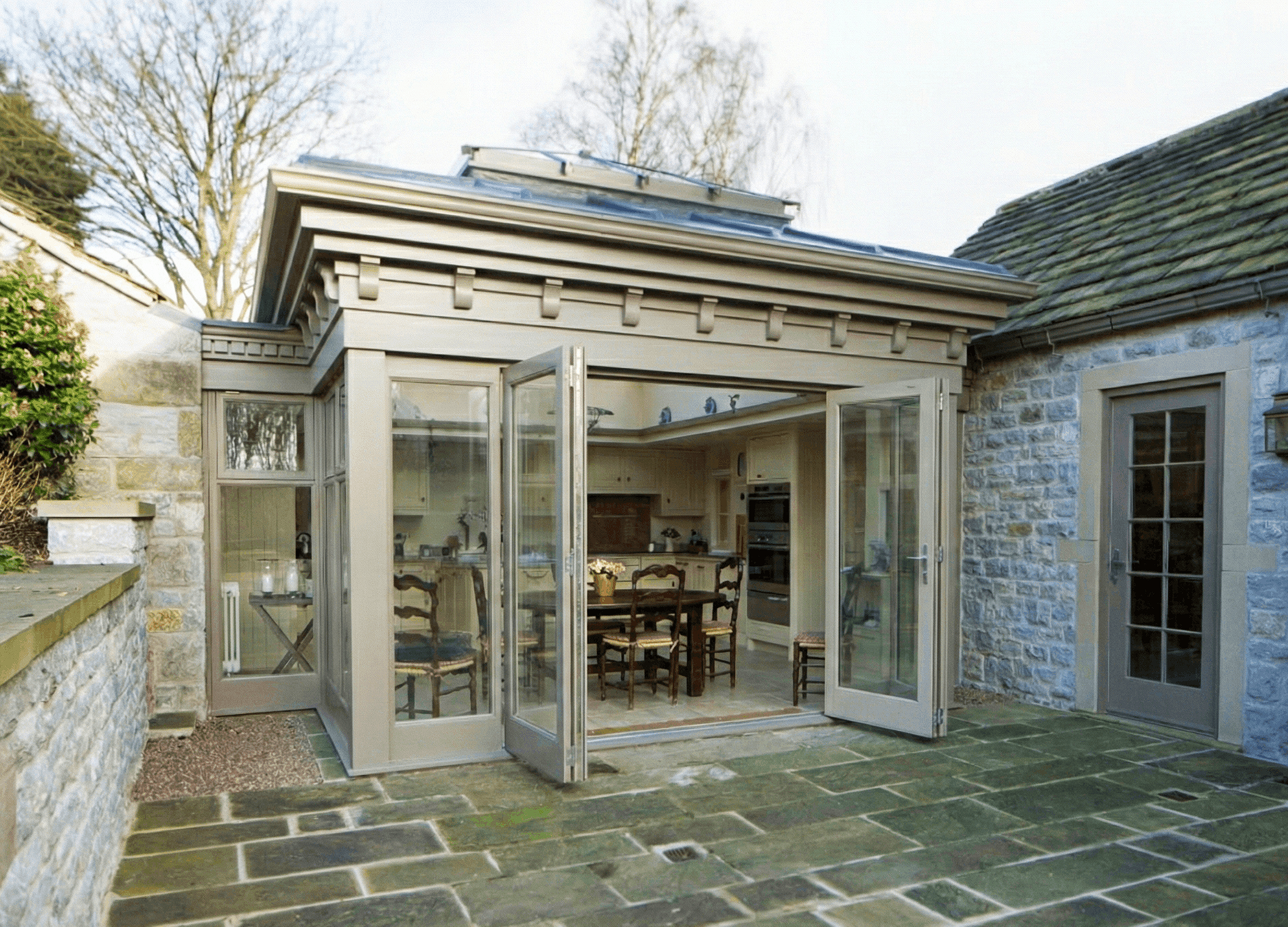 View of a backyard with an outdoor dining area, stone paving, and a building with a glass roof extension, fully open to reveal an interior kitchen and dining space.