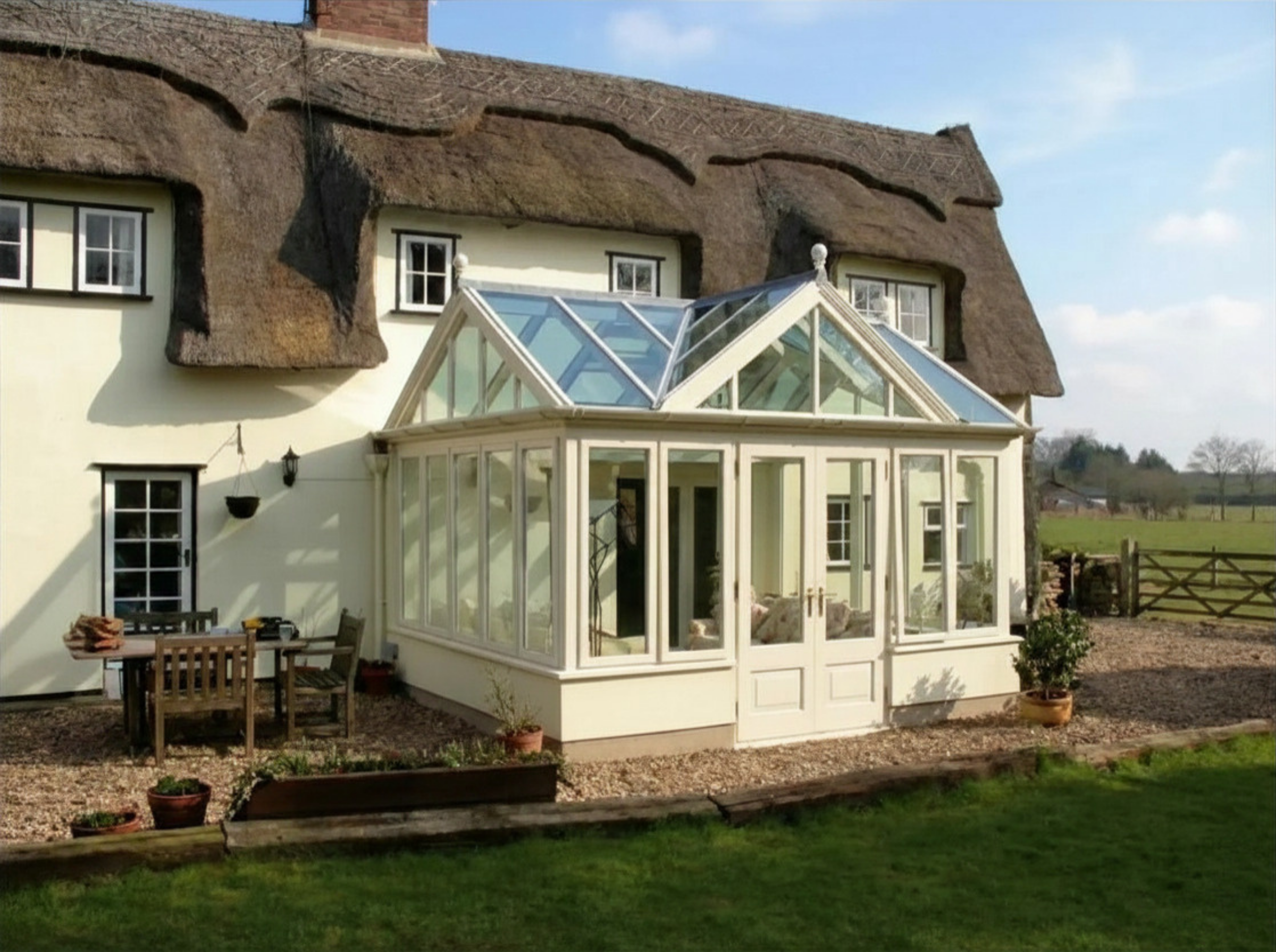 A bespoke cream-painted timber conservatory extension with a pitched glass roof attached to a traditional thatched cottage.