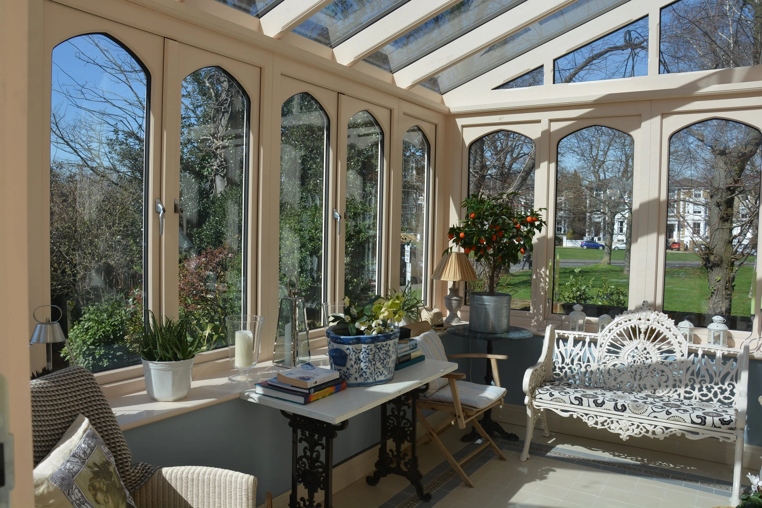 Sunroom with large arched windows and a glass ceiling, decorated with potted plants, a small table with books, a bench with a patterned cushion, and trees with bare branches visible outside.