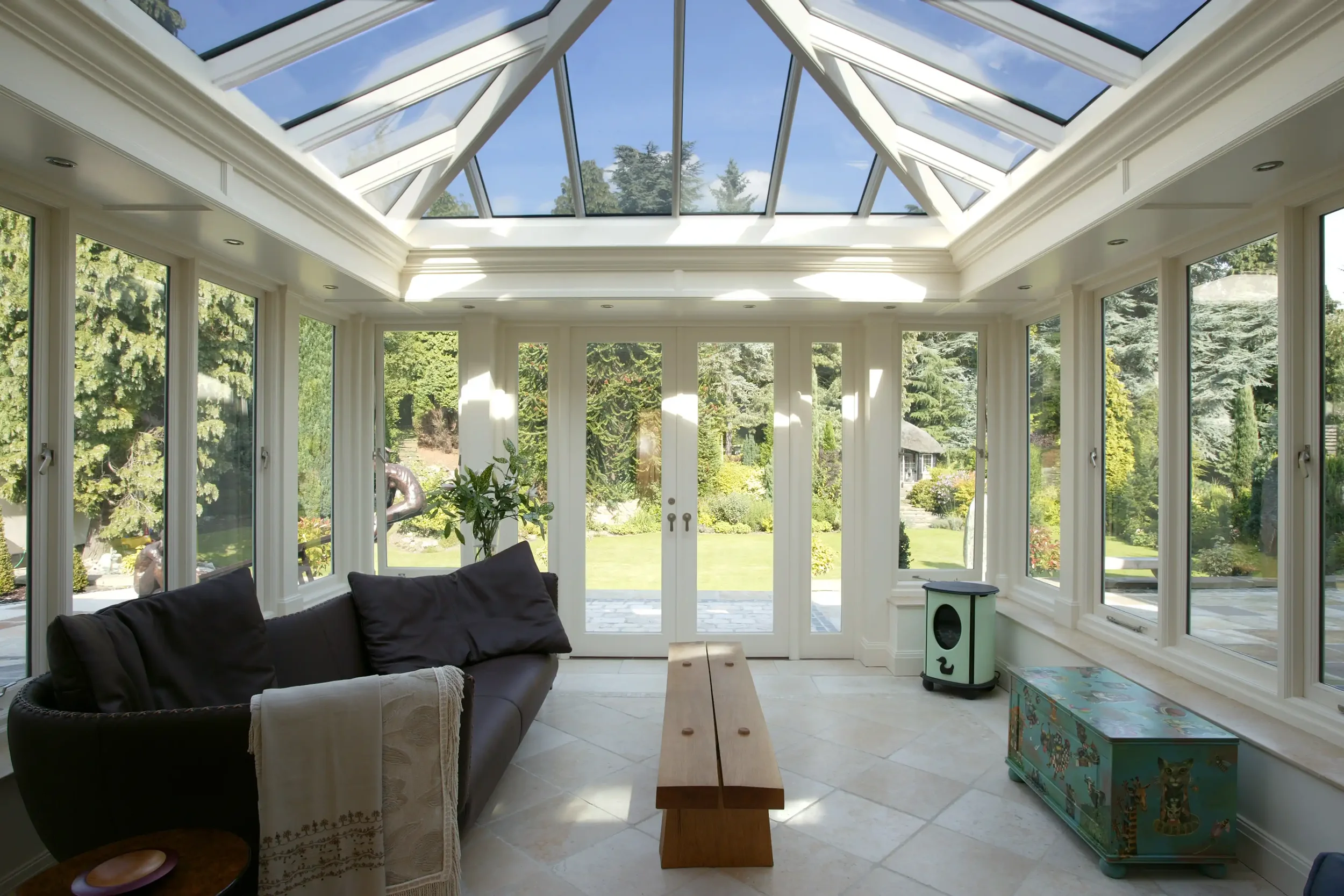 Light-filled timber orangery interior with a large glazed roof lantern, garden-facing doors and natural stone flooring.