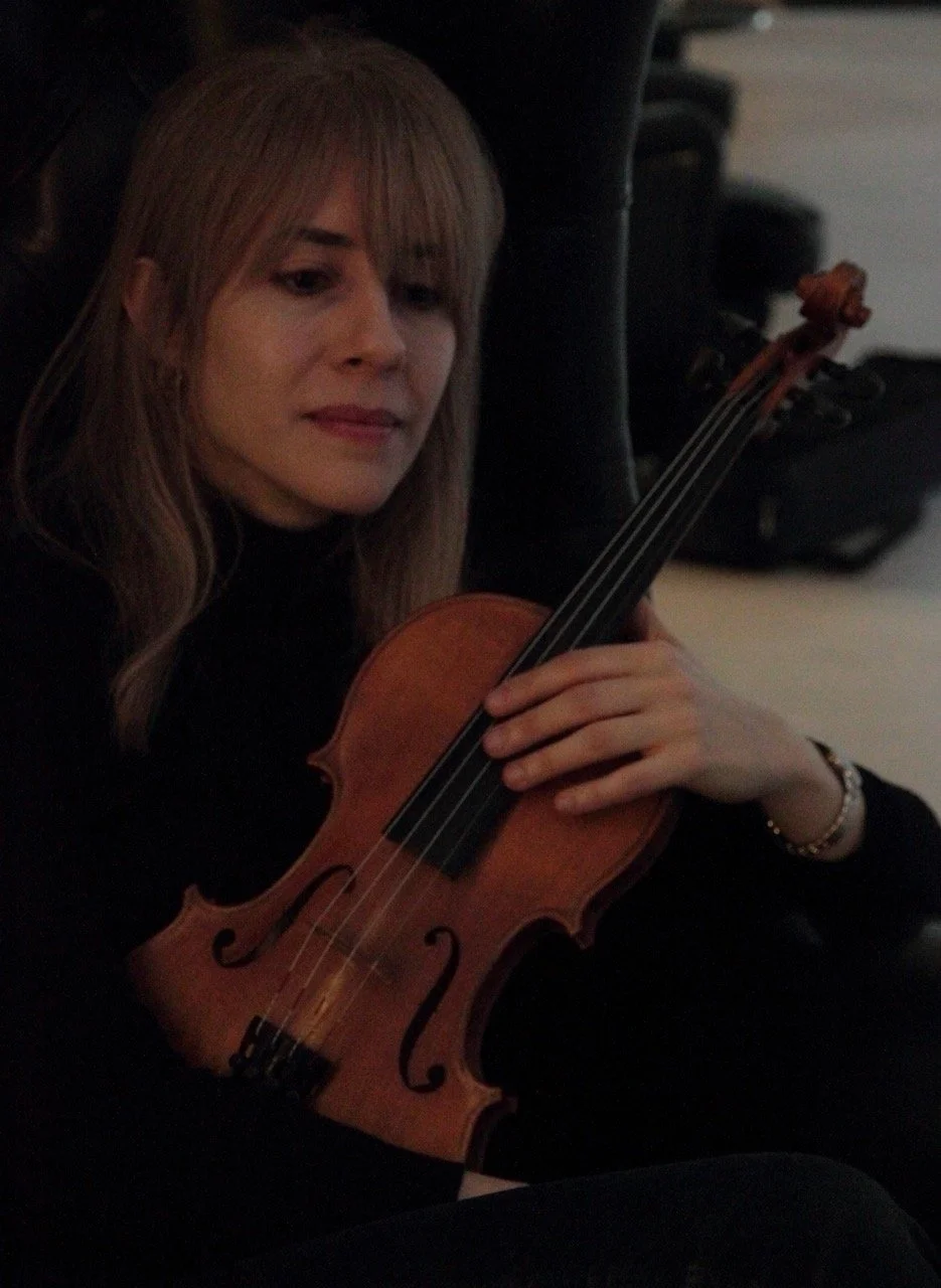 A woman, Talia Ramos, holding her violin in front sitting portrait against a dim lit room background.