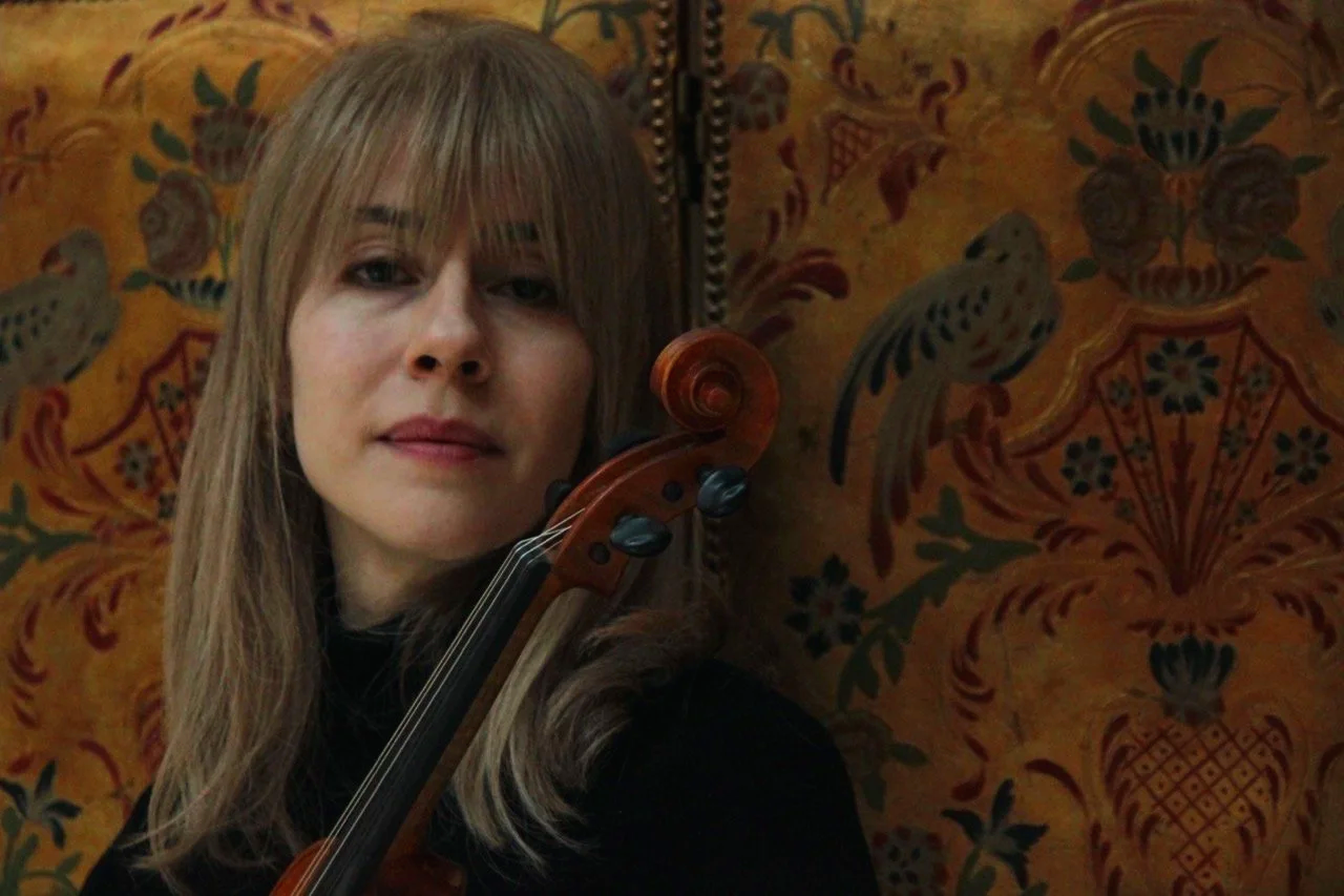 Talia Ramos, with long blonde hair, holds a violin, standing in front of an ornate, patterned background with floral and bird motifs.