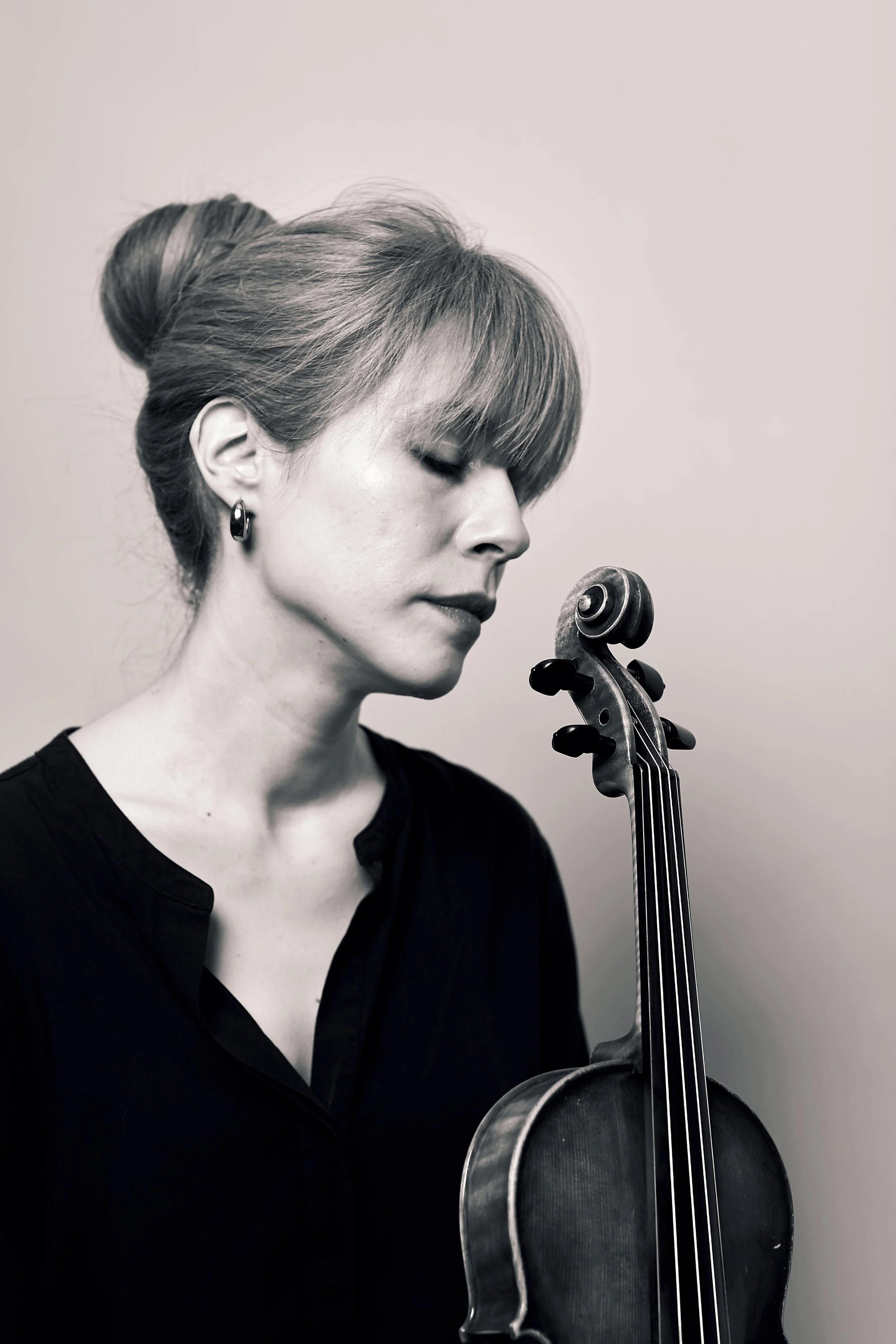 Black and white photo of a woman, Talia Black-and-white photo of a woman, Talia Ramos, with styled hair and earrings, holding a violin and looking down.Ramos, with styled hair and earrings, holding a violin and looking down.