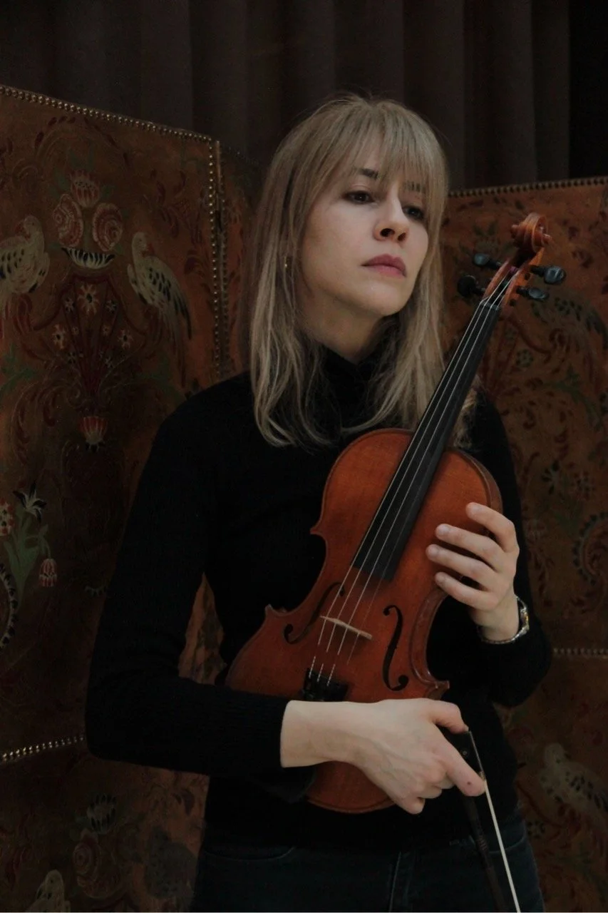 A whole body portrait of a woman, Talia Ramos, holding her violin and bow against a flowery pattern background.