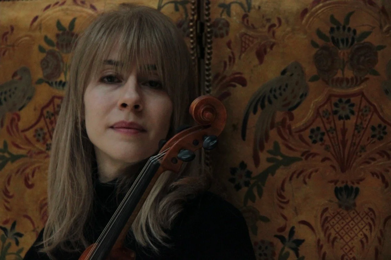 A front profile of a woman, Talia Ramos, holding her violin on the side, gently smiling against an old-fashioned nature pattern background.